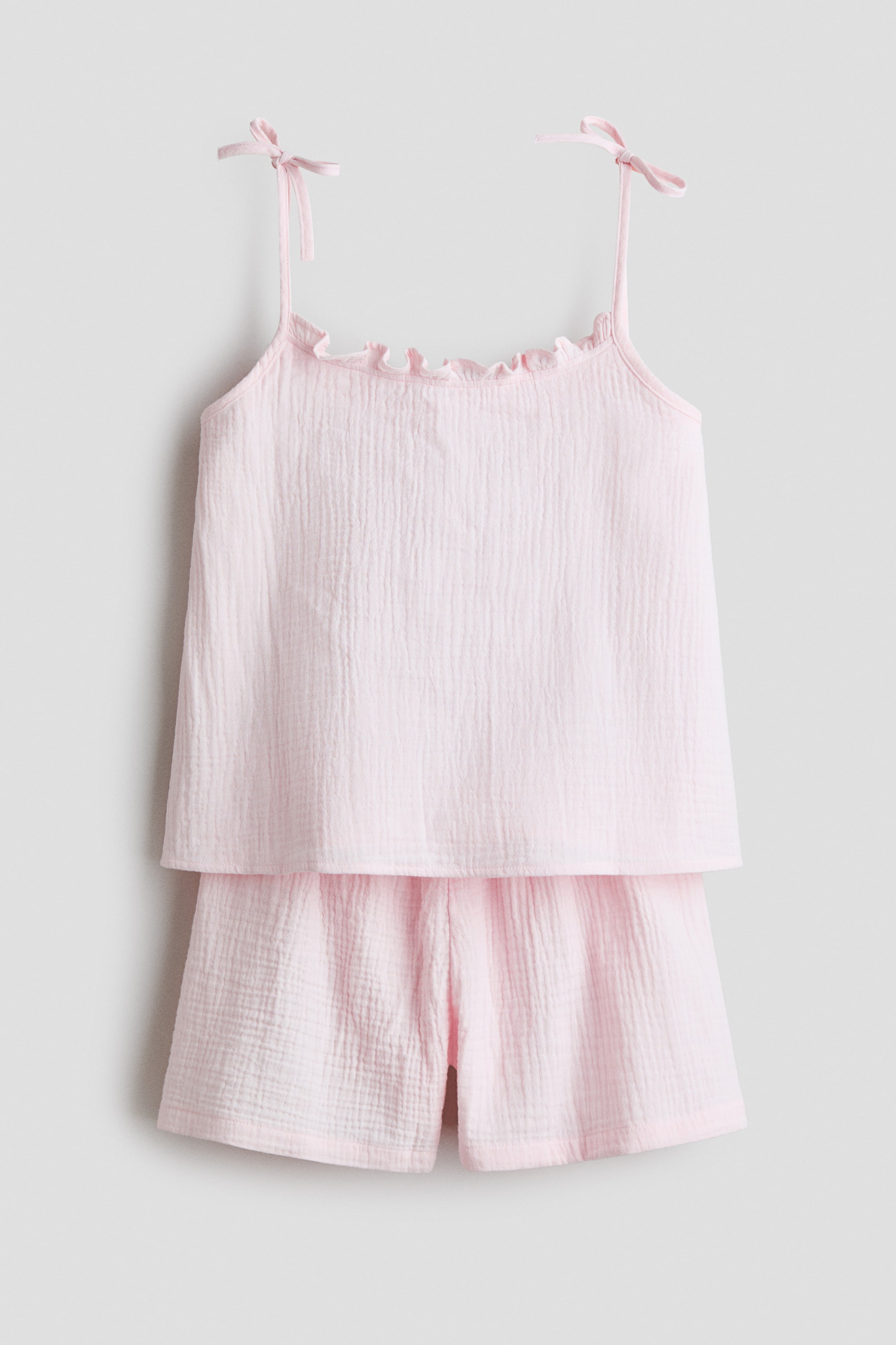 View larger image: A light pink pyjama set is showcased front-facing, comprising a strappy top and shorts in crinkled cotton muslin. The top features a frill-trimmed neckline and narrow shoulder straps detailed with bows, while the shorts display covered elastication at the waist.
