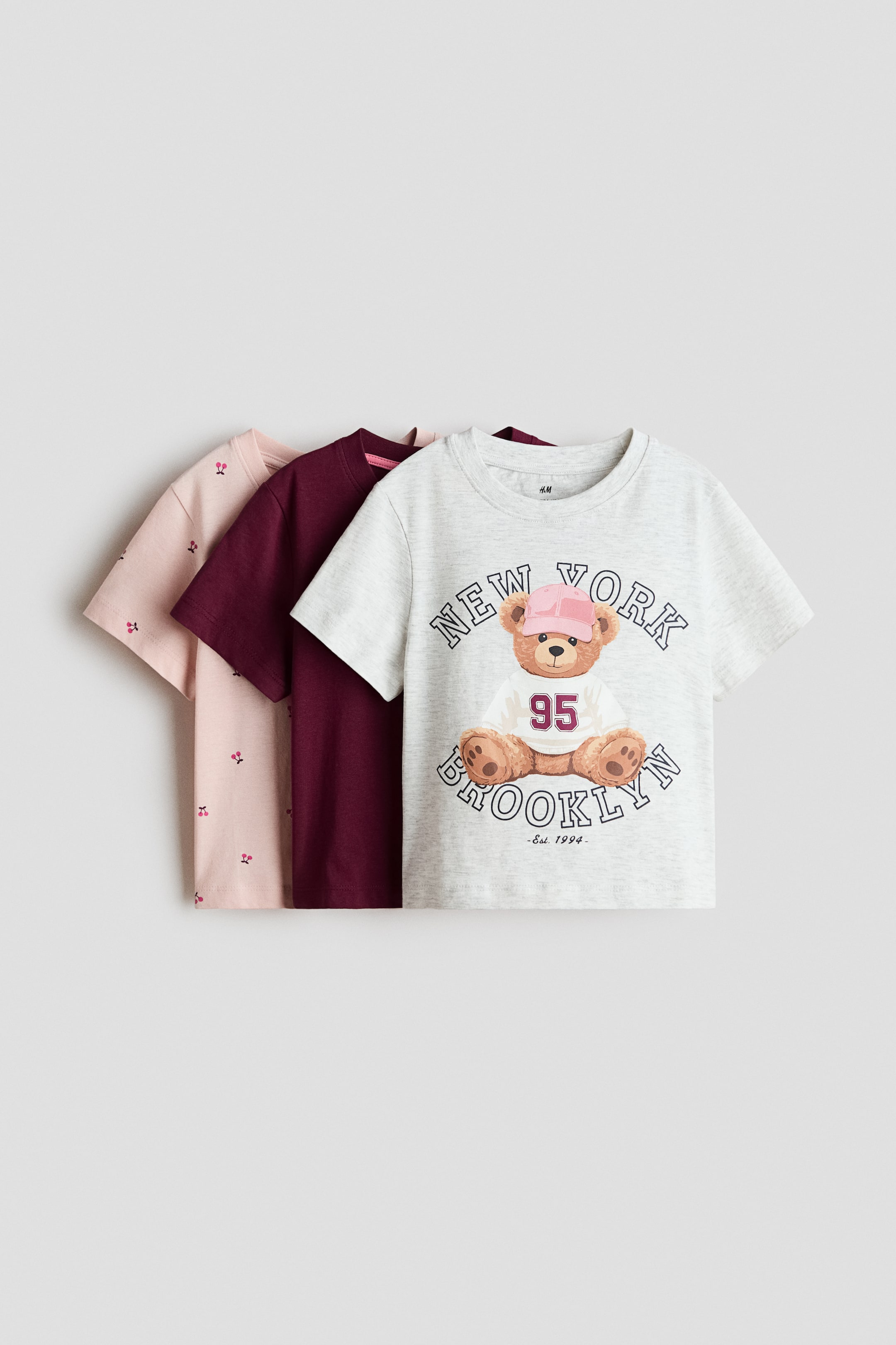 View larger image: Three cotton jersey T-shirts are shown in their front views, layered diagonally. Each one features a ribbed neckline and gently dropped shoulders. The front T-shirt displays a light gray marl fabric with a printed teddy bear wearing a pink cap and the text "NEW YORK BROOKLYN." Behind it, a solid burgundy T-shirt and a light pink T-shirt with small cherry prints are layered.