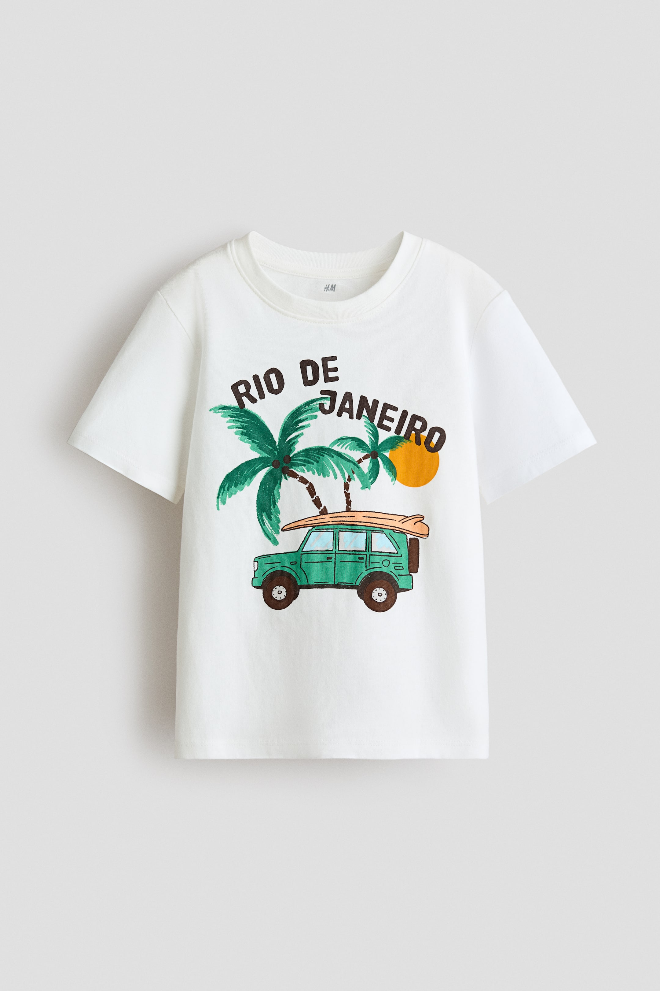 Ver imagen más grande: Una camiseta blanca presenta su parte delantera, con un cuello redondo acanalado, hombros caídos y un dobladillo recto. Un estampado gráfico en el frente muestra el texto 'RIO DE JANEIRO' sobre dos palmeras verdes, un sol naranja y un vehículo todoterreno verde con una tabla de surf en el techo.