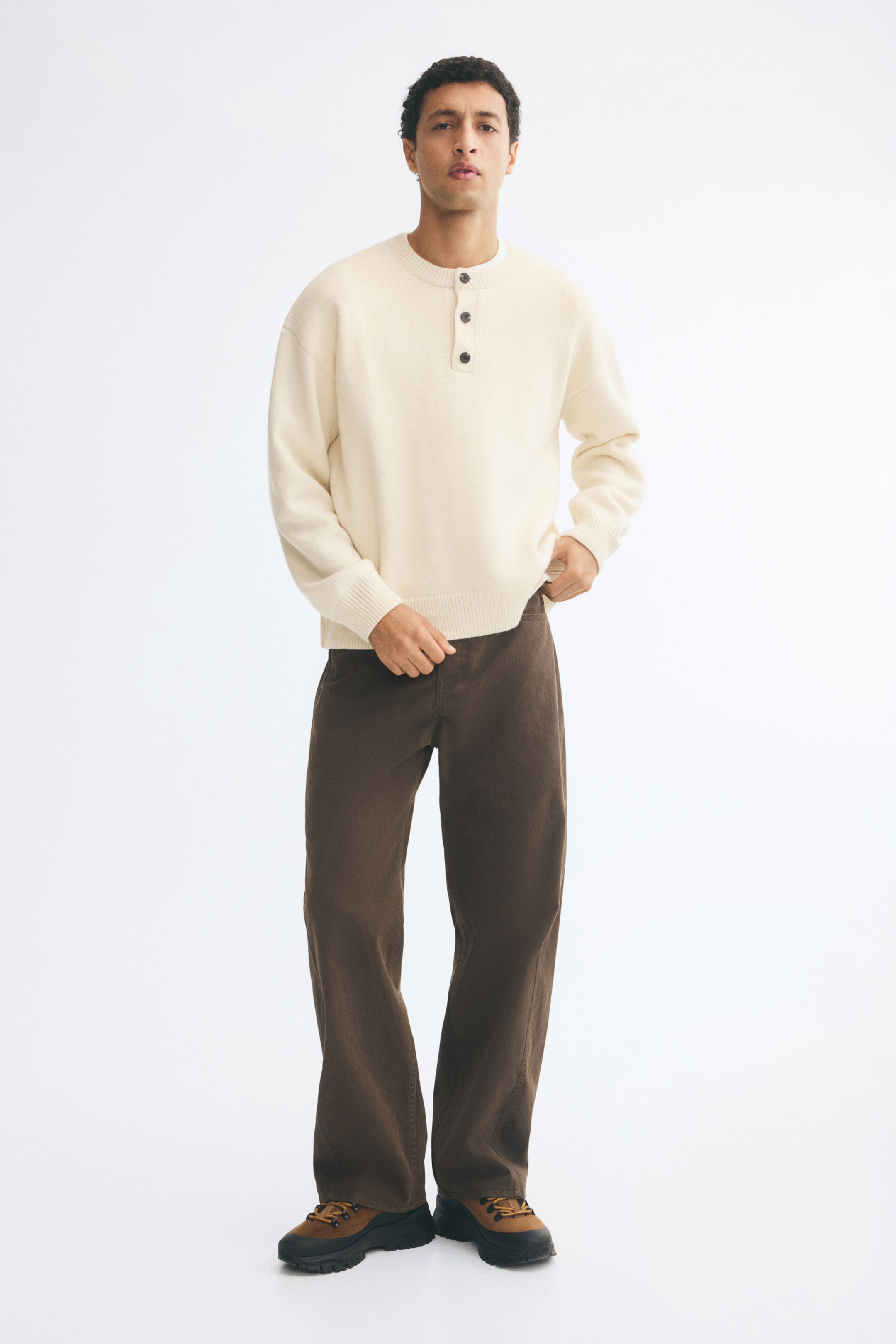 View larger image: Brown cotton twill trousers featuring a relaxed fit, straight legs, and forward-facing side seams are being worn by the model. The trousers are being paired with a cream-coloured knit henley jumper and brown boots.