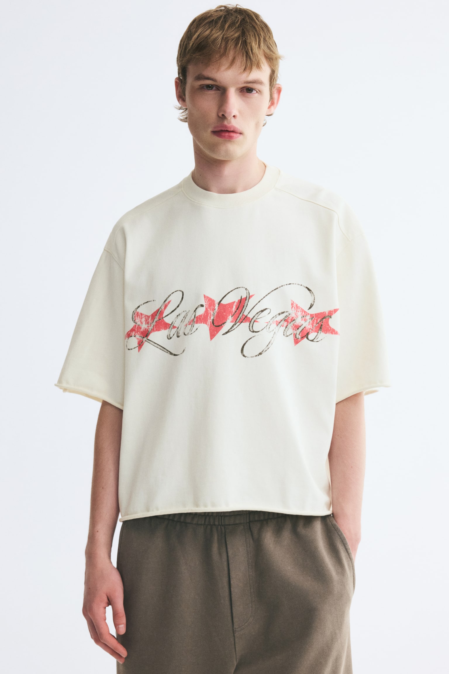 An off-white, boxy-style t-shirt featuring a printed "Las Vegas" graphic across the chest is being worn by the model. The oversized tee is designed with dropped shoulders, a round ribbed neckline, and raw, rolled edges at the cuffs and hem, paired with olive-brown trousers. - 1