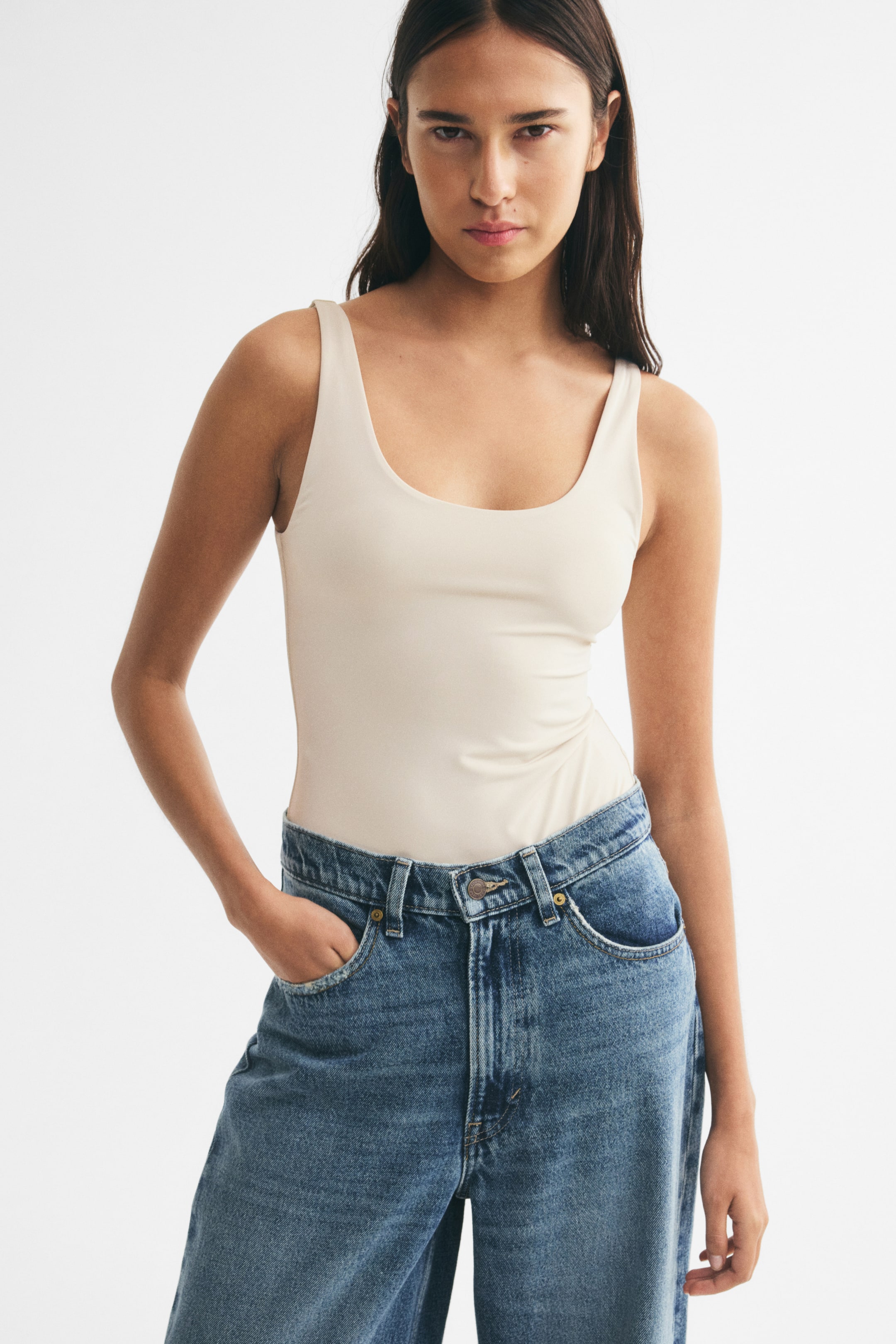 View larger image: A light beige stretch jersey bodysuit with a scoop neckline and wide shoulder straps is worn by the model. The bodysuit is styled tucked into high-waisted blue jeans.