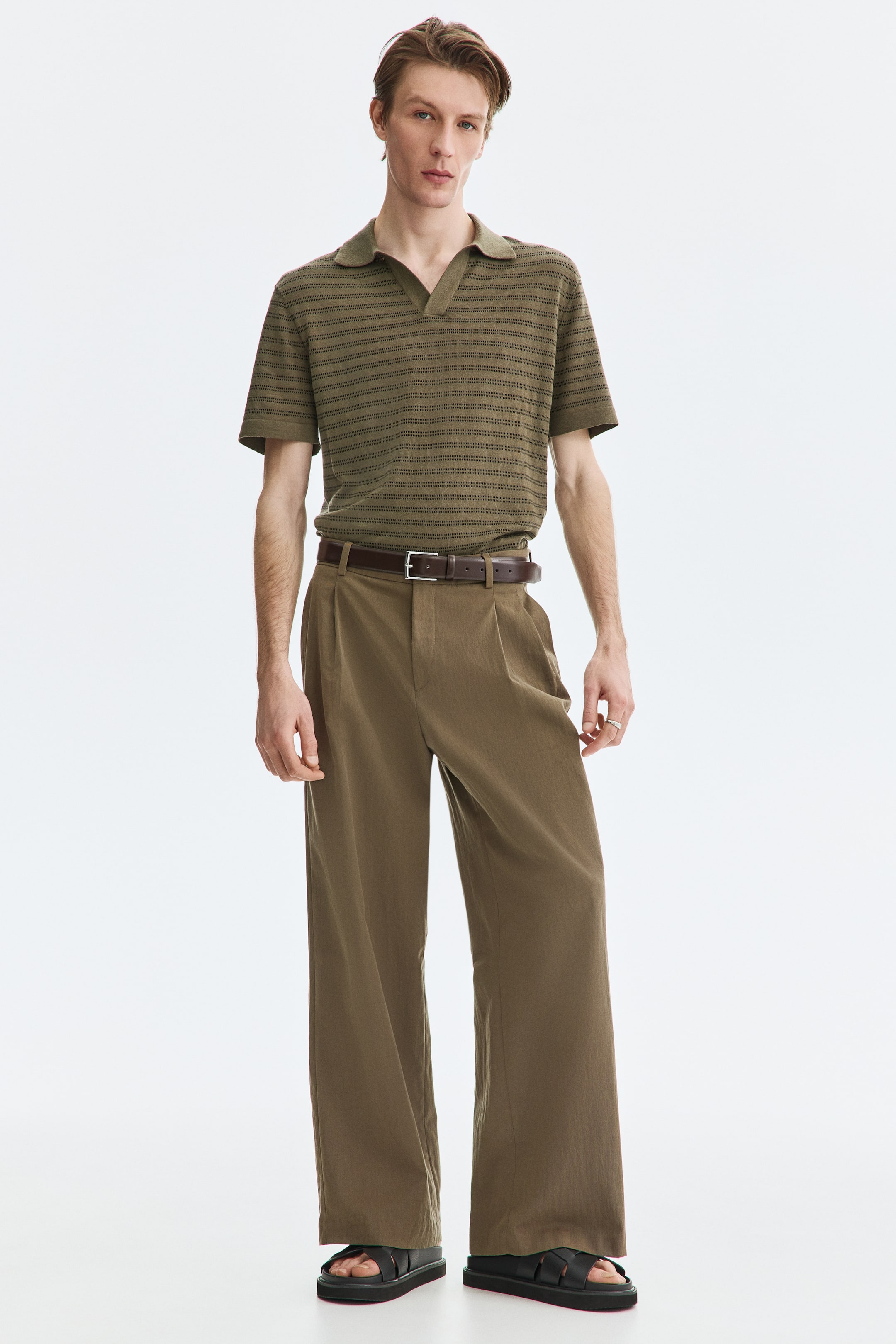 View larger image: The model is wearing an olive green knitted polo shirt adorned with dark horizontal stripes. It features a spread collar and short sleeves, neatly tucked into wide-leg pleated trousers, secured with a dark brown belt.