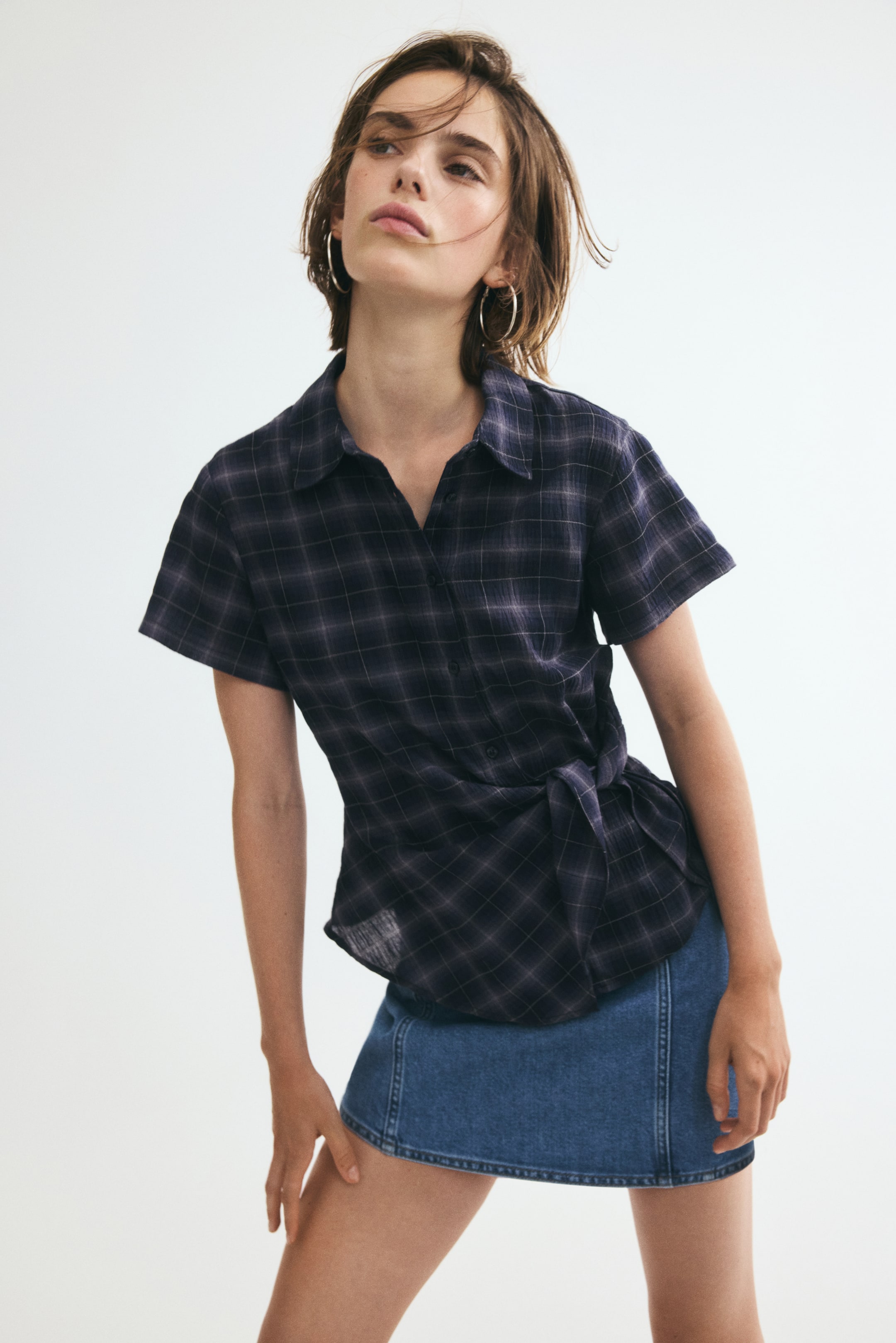 View larger image: The model is wearing a short-sleeved fitted plaid shirt in a smocked weave, featuring a collar, front buttons, and a wide tie detail at one side of the waist. The shirt is paired with a blue denim mini skirt.