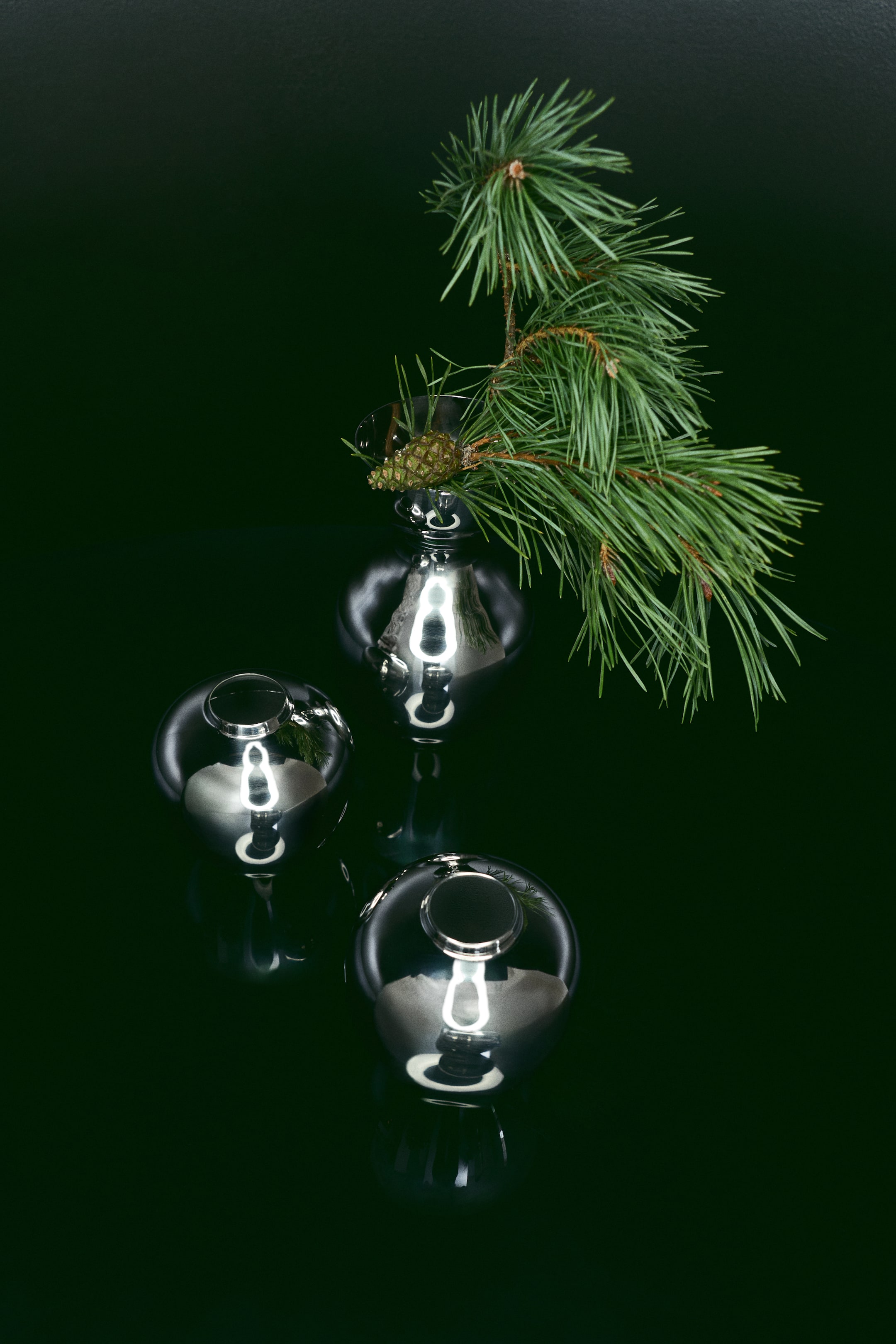View larger image: Three rounded mini vases in highly polished stainless steel reflect light from a high-angle view, displaying their bulbous shapes and narrow openings. One vase holds a green pine branch with a small pine cone, while the other two sit empty in the foreground to showcase their metallic finish.
