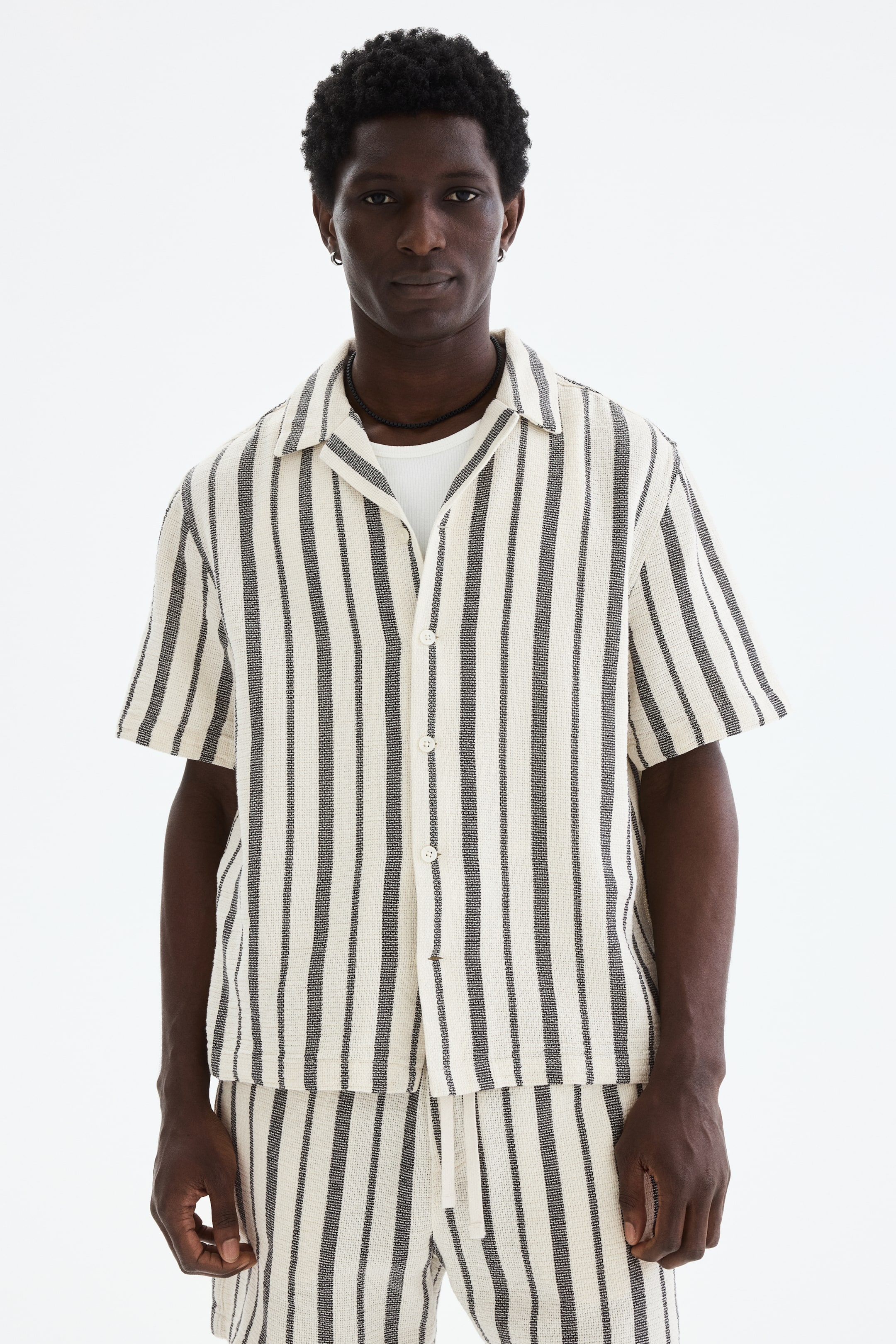 View larger image: A cream-textured short-sleeve shirt with dark vertical stripes is shown from the front, featuring a camp collar and a partially unbuttoned front with light-colored buttons. It is layered over a white T-shirt and paired with matching striped shorts.