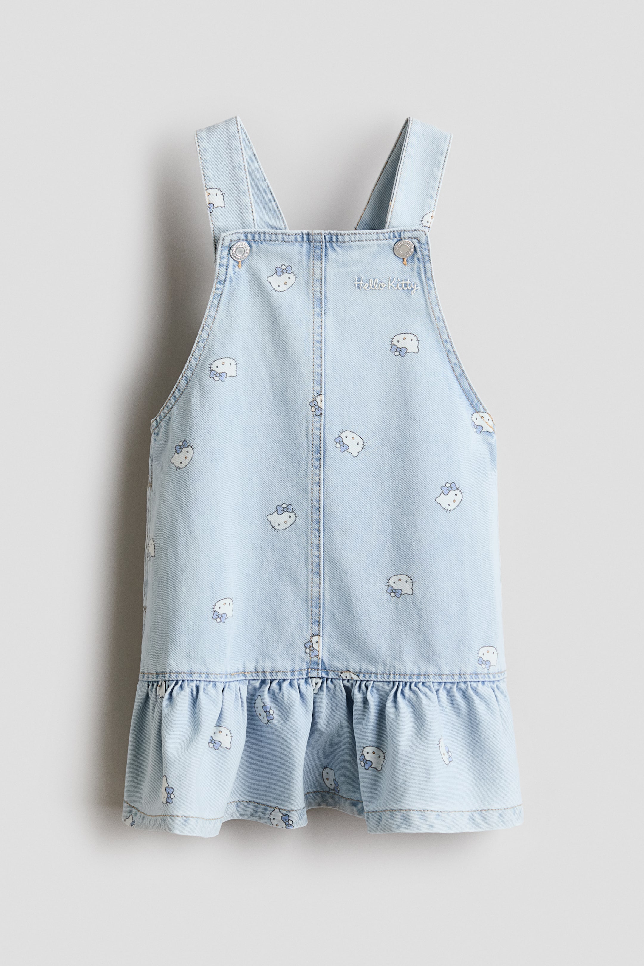 Ampliar la imagen: Un vestido overol de mezclilla azul claro está de frente. Presenta un estampado completo de caras de Hello Kitty con moños, un peto frontal con tirantes ajustables asegurados por botones plateados, texto bordado "Hello Kitty" y un volante en el borde inferior.
