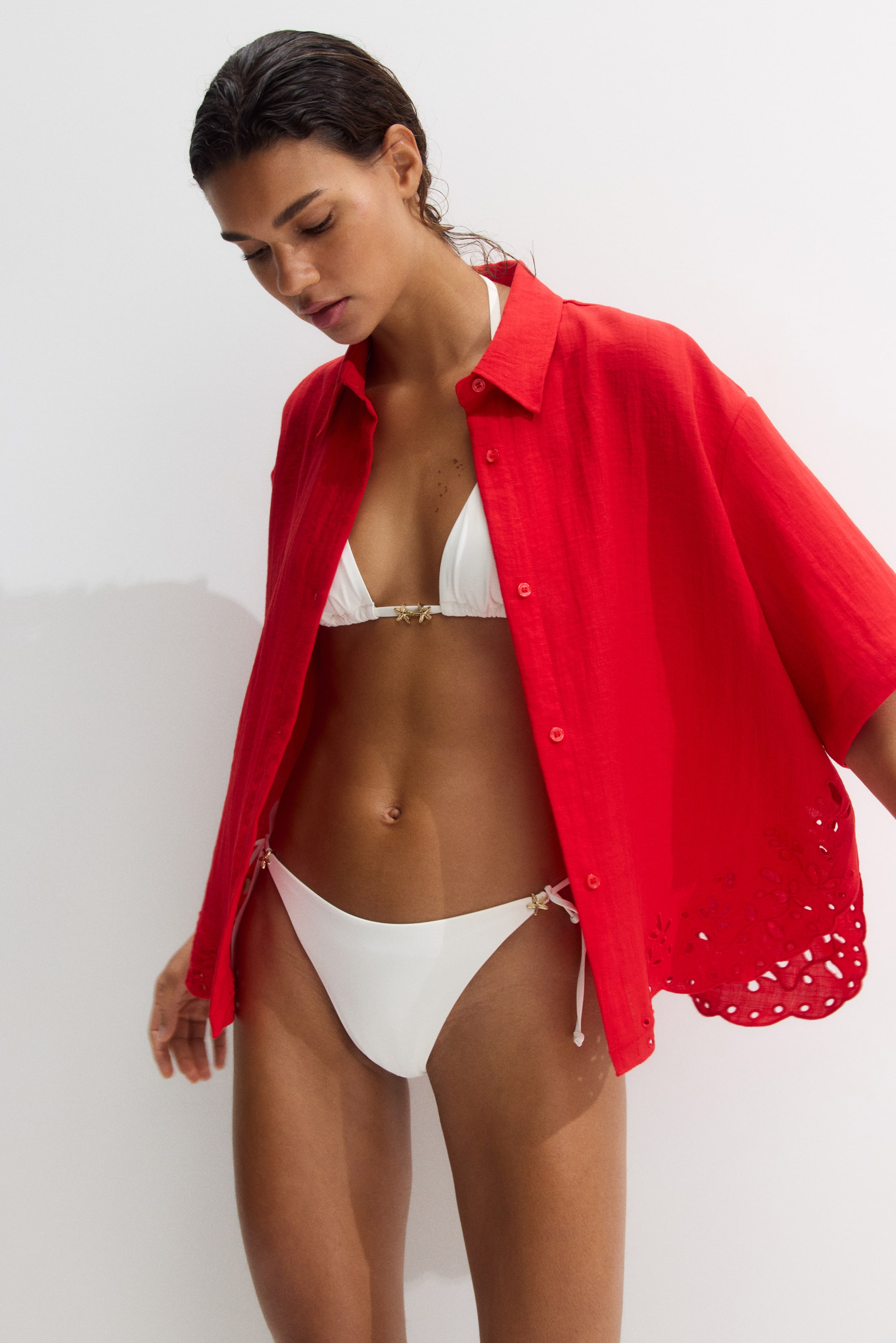 View larger image: The model is wearing a red loose-fit beach shirt open over a white bikini. The woven shirt boasts a collar, dropped shoulders, elbow-length sleeves, and a curved hem adorned with broderie anglaise.