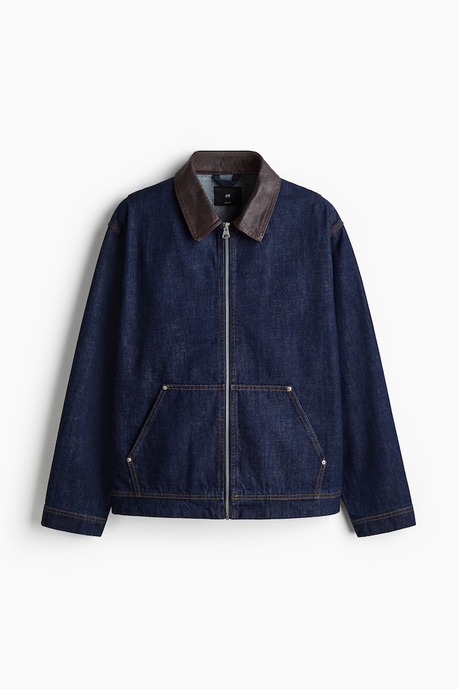 Eine dunkelblaue Jeansjacke wird VORNE angezeigt, mit einem kontrastierenden braunen Kragen und einem silberfarbenen Reißverschluss in voller Länge in der Mitte. Sie ist mit zwei großen, schrägen Aufsatztaschen auf der unteren Vorderseite gestaltet, die jeweils mit zwei silbernen Nieten akzentuiert sind, und kontrastierende braune Nähte sind überall sichtbar. - 1