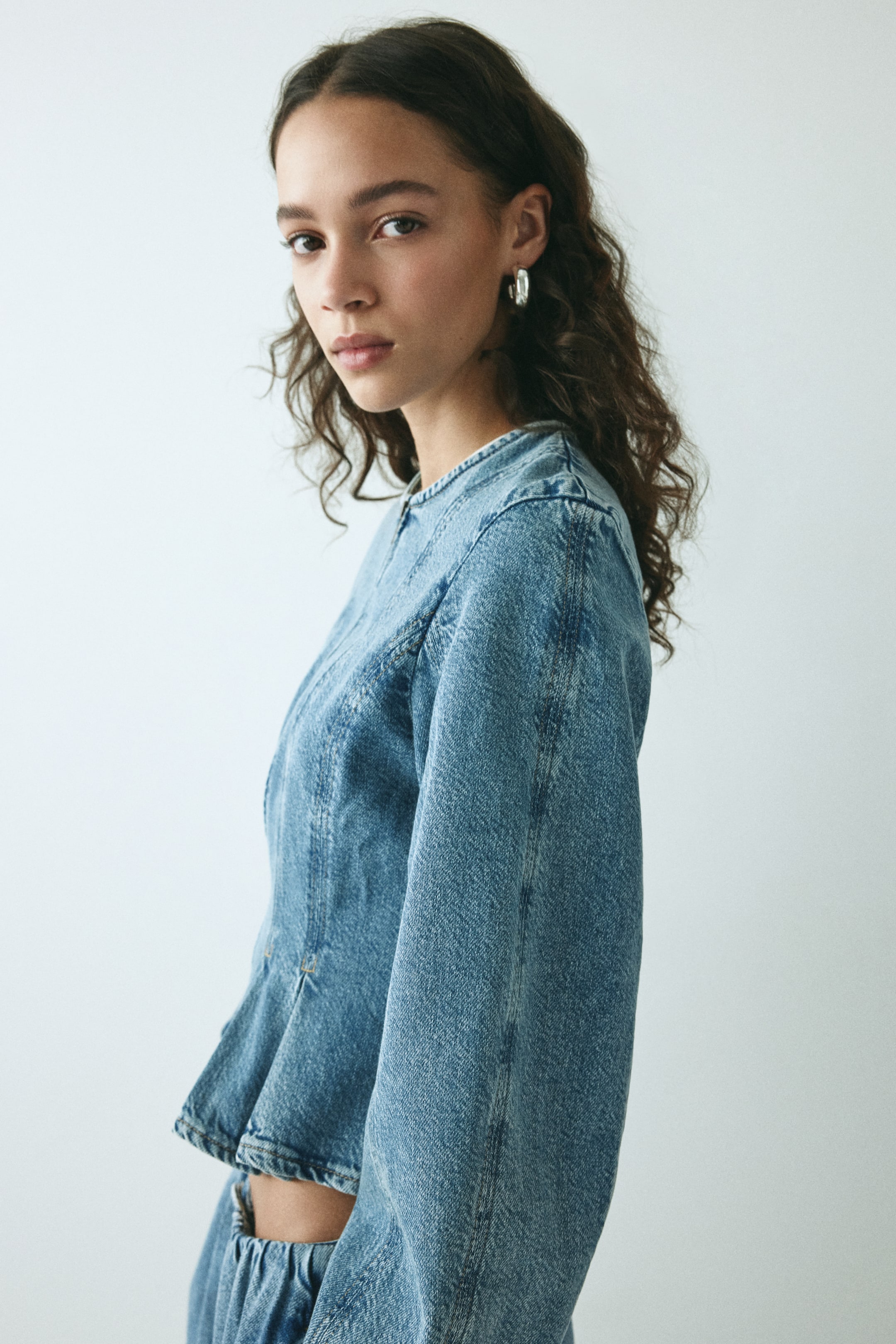 View larger image: A light blue denim top features a fitted bodice with prominent vertical seams, shown from a slight side angle. It displays a flared peplum hem, long sleeves, and a collarless neckline, with matching denim bottoms visible below its cropped length.