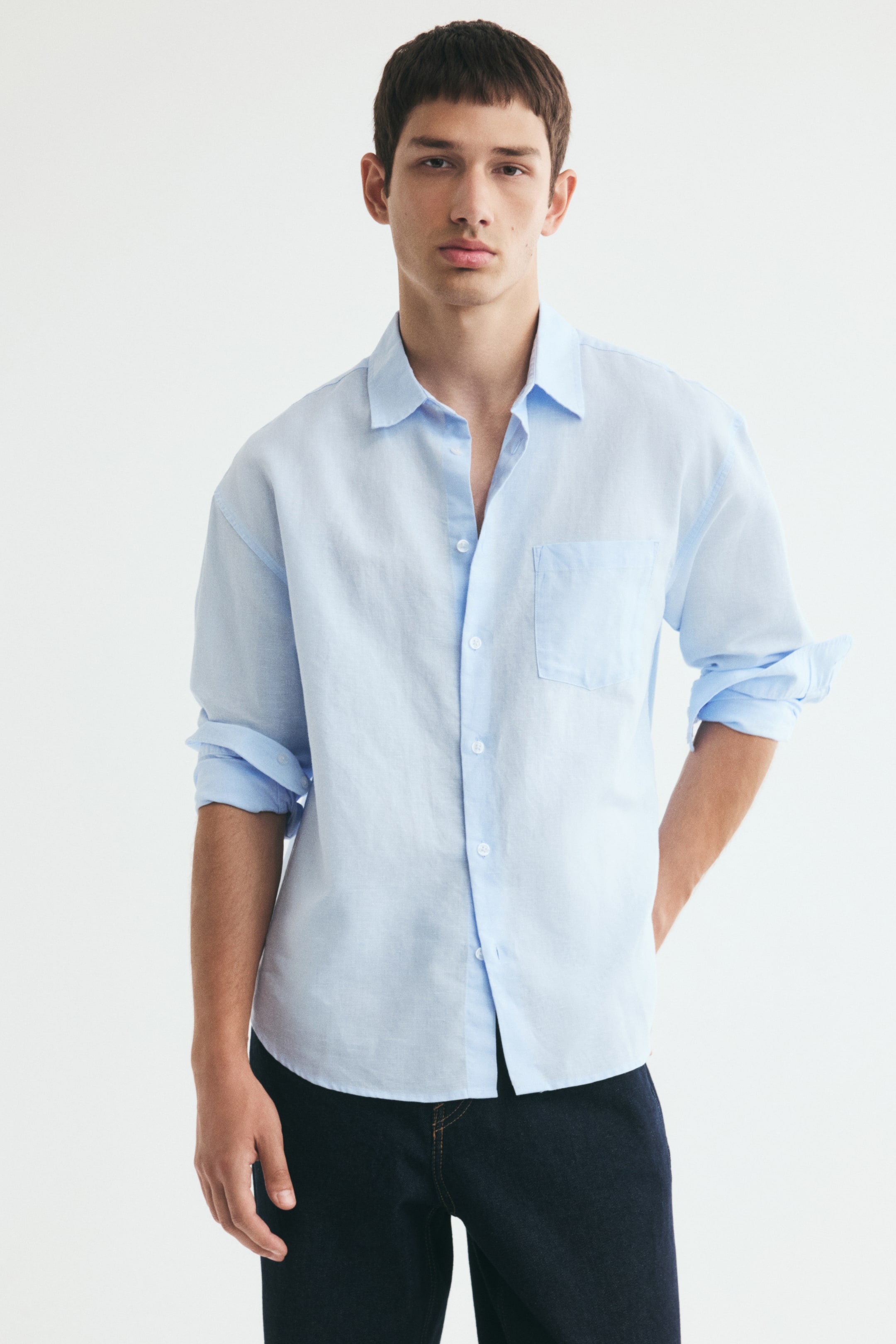 View larger image: A light blue shirt in a textured cotton and linen weave is worn by the model with the top buttons undone and the long sleeves rolled up. The relaxed-fit shirt features a turn-down collar, a single chest pocket, and a rounded hem, and is styled untucked over dark blue jeans.