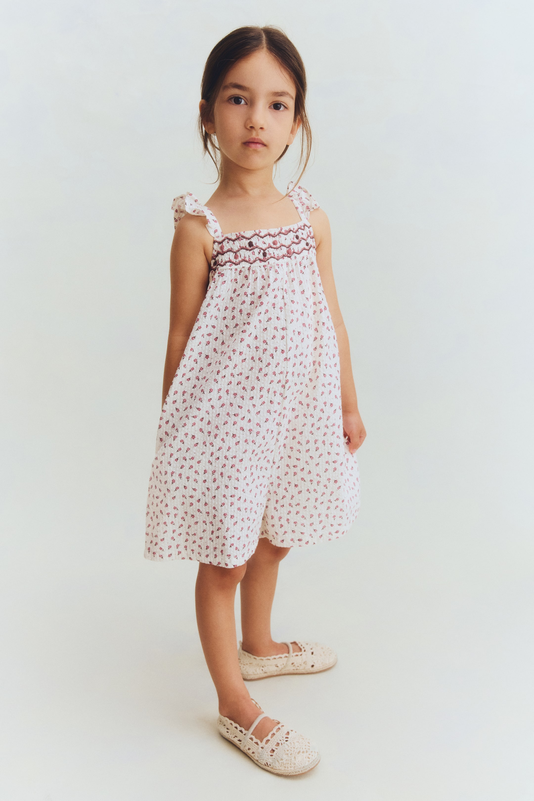 View larger image: A white dress, showcasing a small, repeating reddish-pink floral pattern, ruffled shoulder straps, and a dark reddish-brown smocked bodice, is being worn. It is paired with light cream crocheted flat shoes, featuring an intricate openwork design and a single instep strap.
