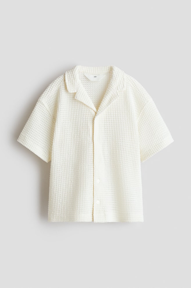 Ein cremefarbenes Shirt im Waffelstrick präsentiert seine Vorderseite, mit einem Reverskragen, kurzen Ärmeln und einem Knopfverschluss vorne mit hellen Knöpfen. - 1