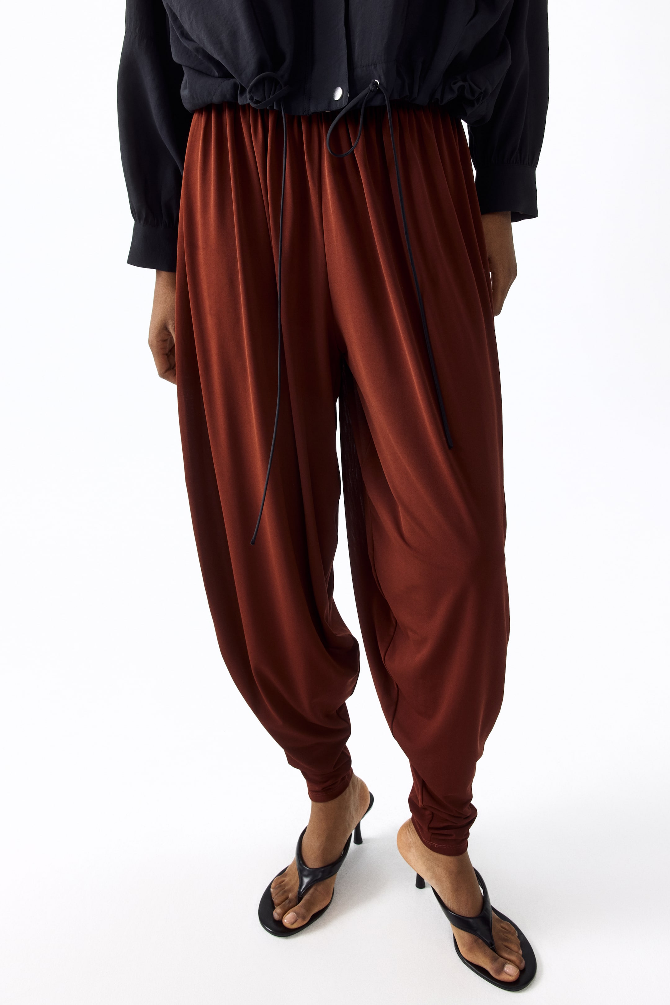 Vis større bilde: De rustfargede buksene har en voluminøs, avslappet passform, som samler seg ved anklene. En snor i livet med synlige svarte bånd strammer toppen av buksene.