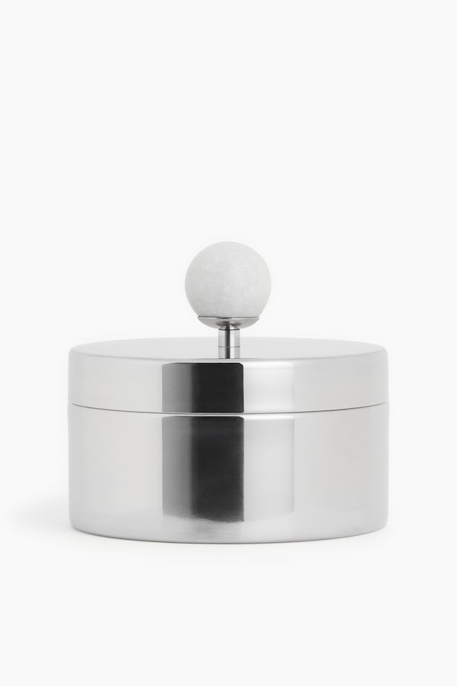 Un conteneur en métal poli de couleur argentée est présenté de face. Il se caractérise par un corps cylindrique et un couvercle plat, avec un bouton sphérique en marbre blanc s'élevant au centre du couvercle sur une tige métallique élancée. - 1