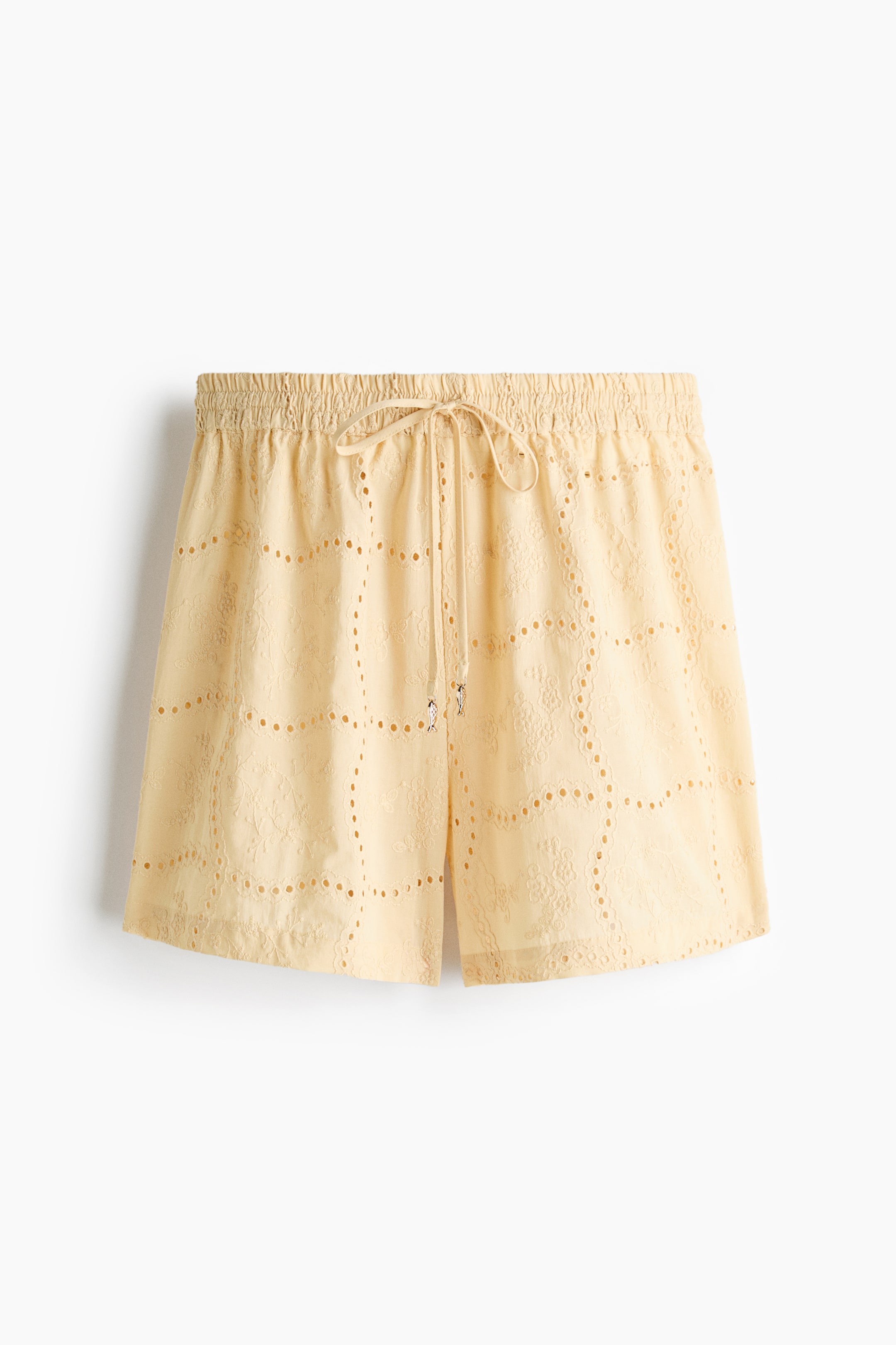 View larger image: The light-yellow shorts are shown from the front, featuring an elasticated, shirred waistband and a matching drawstring tied into a bow at the centre front, with two small metallic charms. The shorts are crafted from broderie anglaise fabric, featuring a repeating, grid-like pattern of cut-out squares with floral motifs, edged by smaller cut-out circles.