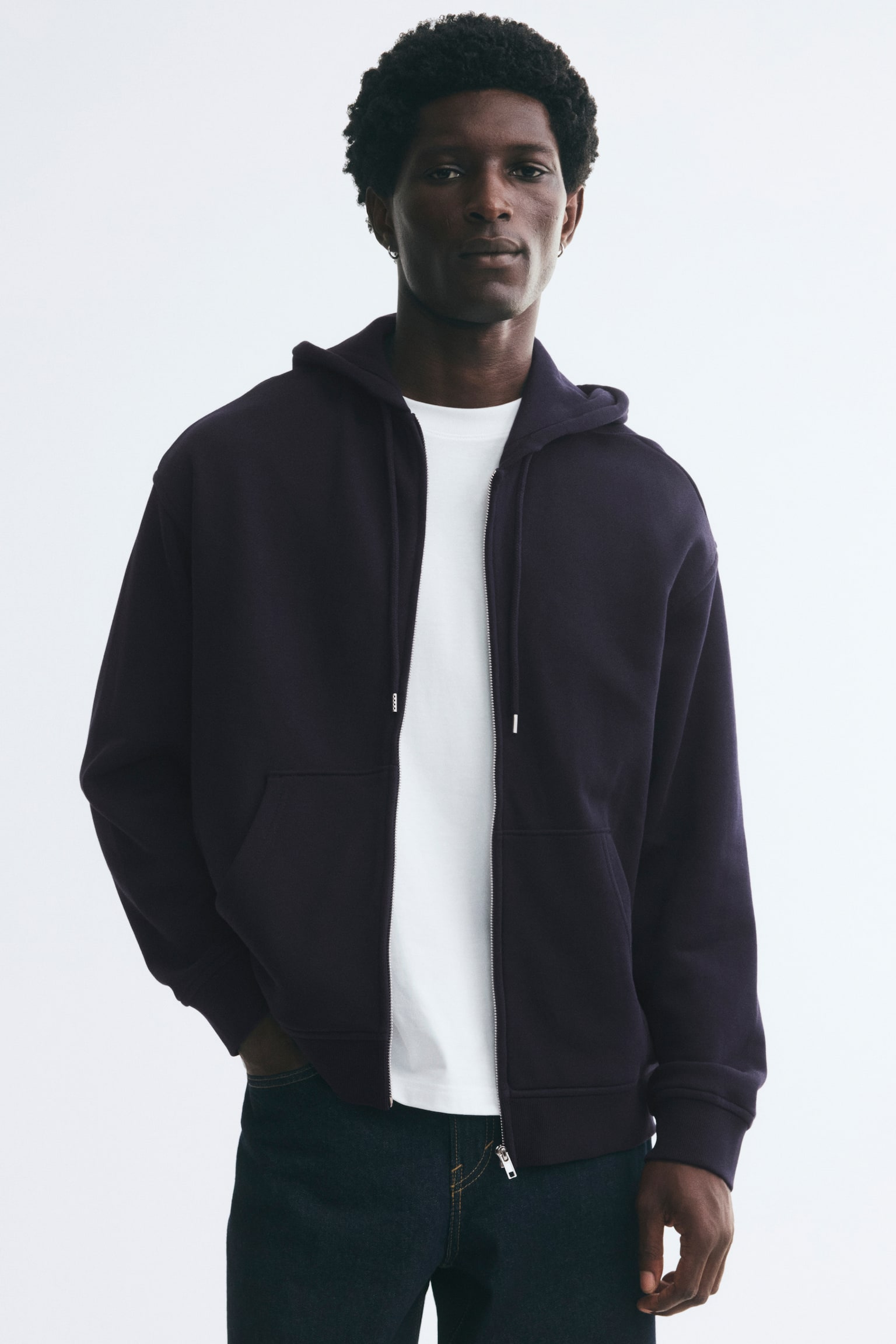 A dark purple zip-up hoodie is worn open by the model over a plain white t-shirt and dark jeans. The hoodie features a relaxed fit, a drawstring hood, dropped shoulders, front pockets, and ribbed cuffs and hem. - 1