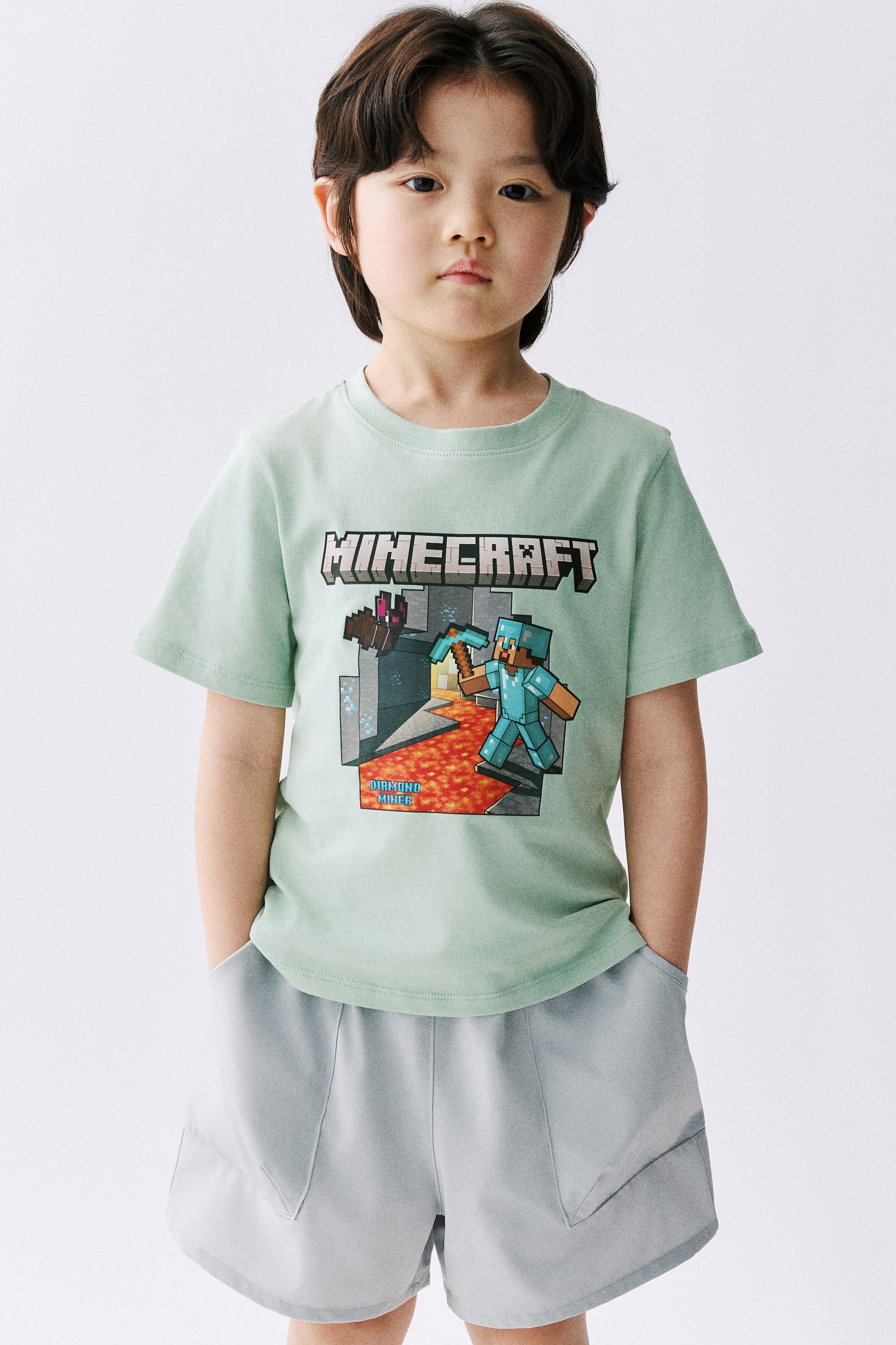 이미지 확대 보기: 연한 녹색 반소매 티셔츠는 앞면에 'Minecraft' 그래픽이 인물과 텍스트와 함께 표시되어 있습니다. 이 티셔츠는 크루넥과 여유로운 핏을 특징으로 하며, 사선 앞주머니와 신축성 있는 허리가 있는 연한 회청색 반바지와 매치되어 있습니다.