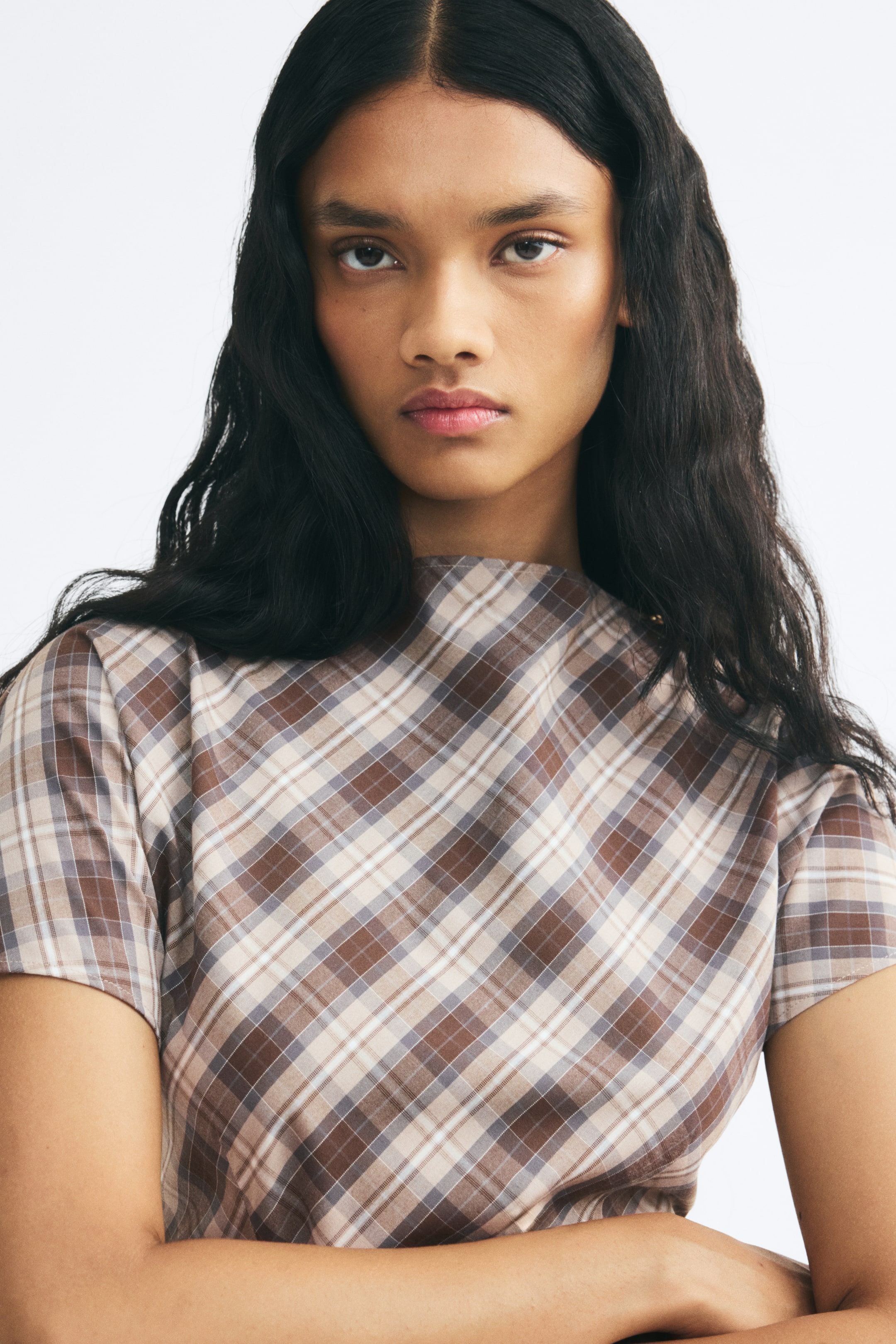 View larger image: A fitted, short-sleeved top with a brown and beige plaid pattern is worn by the model. The top features a high boat neck and a diagonal check fabric design.