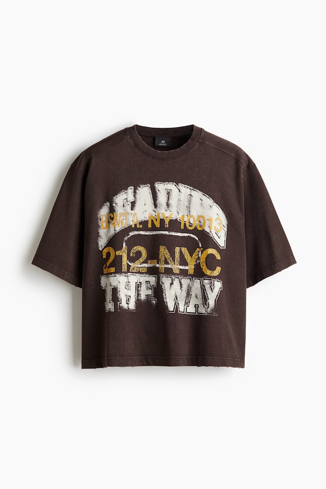A dark brown, short-sleeved t-shirt is being presented from the front, featuring a prominent distressed graphic print. The print displays layered text, including "LEADING" and "THE WAY" in white, and "B'KLYN NY 10013" and "212-NYC" in gold, with the "212-NYC" text enclosed in an outline, all set against the t-shirt's crew neck and raw-edge hem. - 1