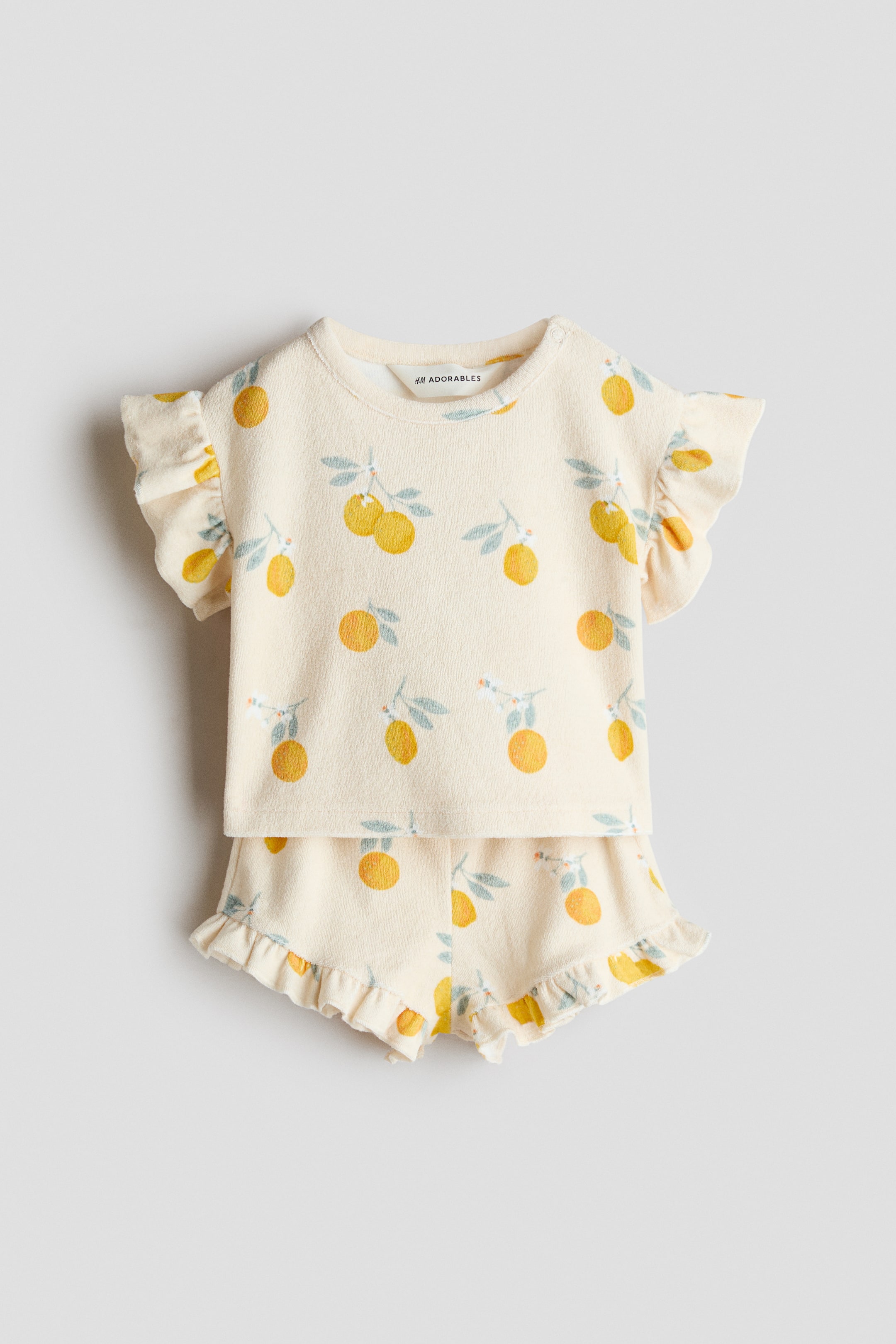 Agrandir l'image: Un ensemble deux pièces pour bébé comprend un haut de couleur crème et un short assorti, tous deux ornés d'un imprimé répétitif de fruits d'agrumes jaunes et oranges avec des feuilles vertes et des fleurs blanches. Le haut à manches courtes présente des détails à volants sur les manches et un col rond, tandis que le short arbore des ourlets à volants.