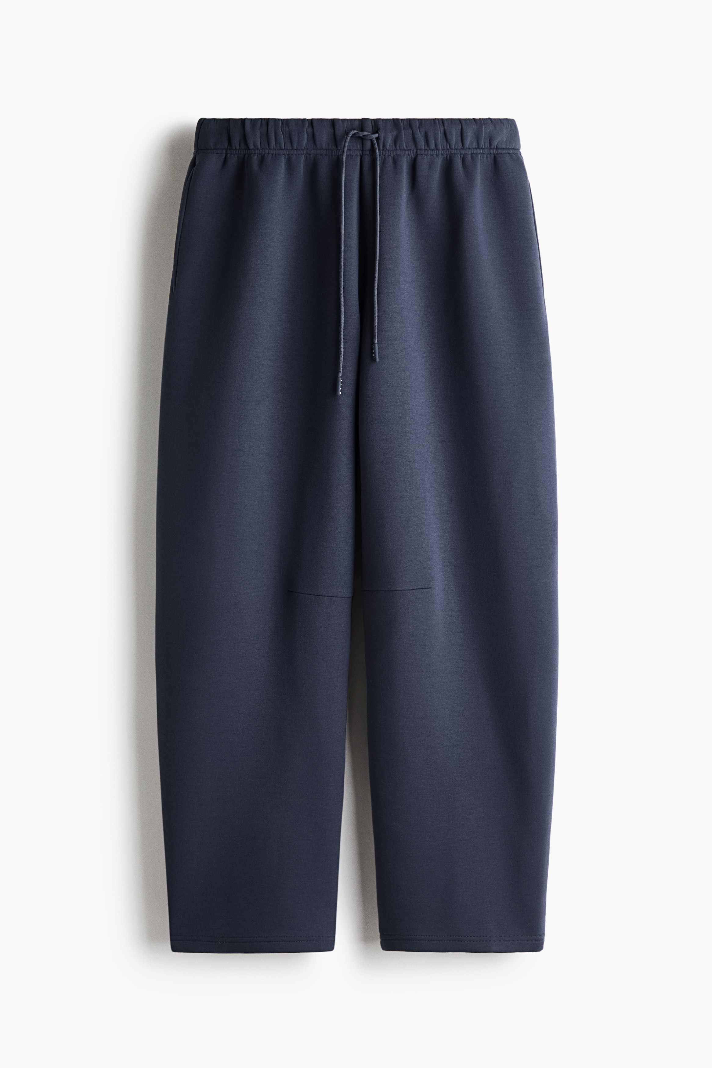 A light grey crew-neck T-shirt with an oversized fit and dark blue wide-leg sweatpants are being worn by the model. The model is standing with hands placed in the sweatpants' pockets, completing the outfit with grey and white trainers.