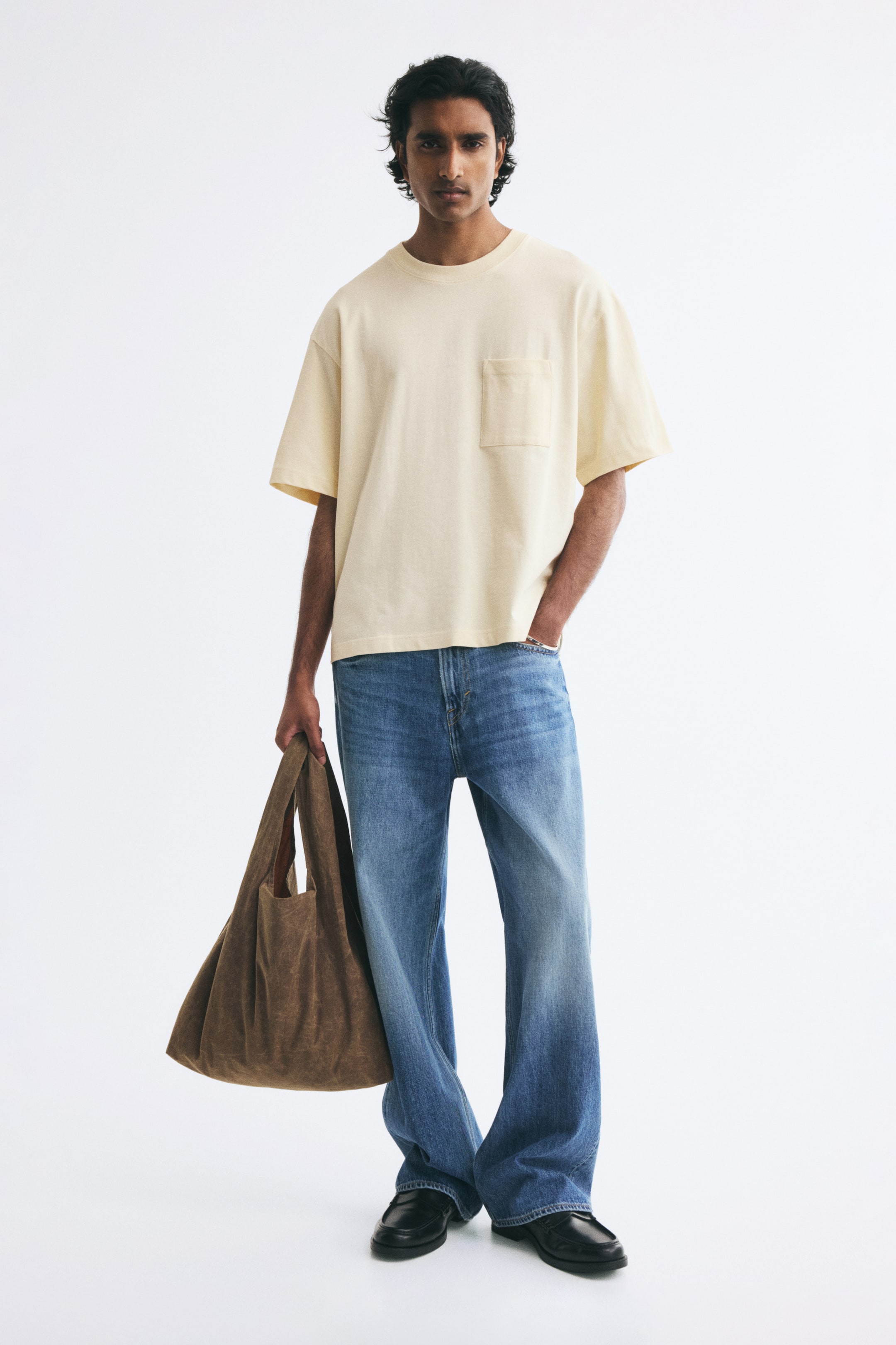 View larger image: A light yellow, heavyweight cotton T-shirt featuring a relaxed fit, a rib-trimmed round neckline, dropped shoulders, a straight hem, and a single chest pocket is worn by the model. The model styles the tee with loose-fit blue jeans while holding a brown tote bag.