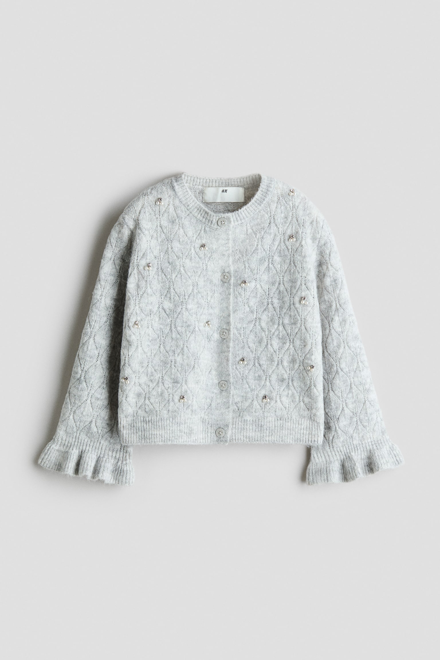 Eine hellgraue Strickjacke ist von vorne zu sehen und verfÃ¼gt Ã¼ber eine strukturierte Strickoptik im Rautenmuster, die mit vereinzelt angebrachten kleinen Kunstperlen verziert ist. Das Design umfasst eine durchgehende Knopfleiste mit grauen KnÃ¶pfen, einen gerippten Rundhalsausschnitt sowie lange Ãrmel mit ausgestellten, gerÃ¼schten BÃ¼ndchen.