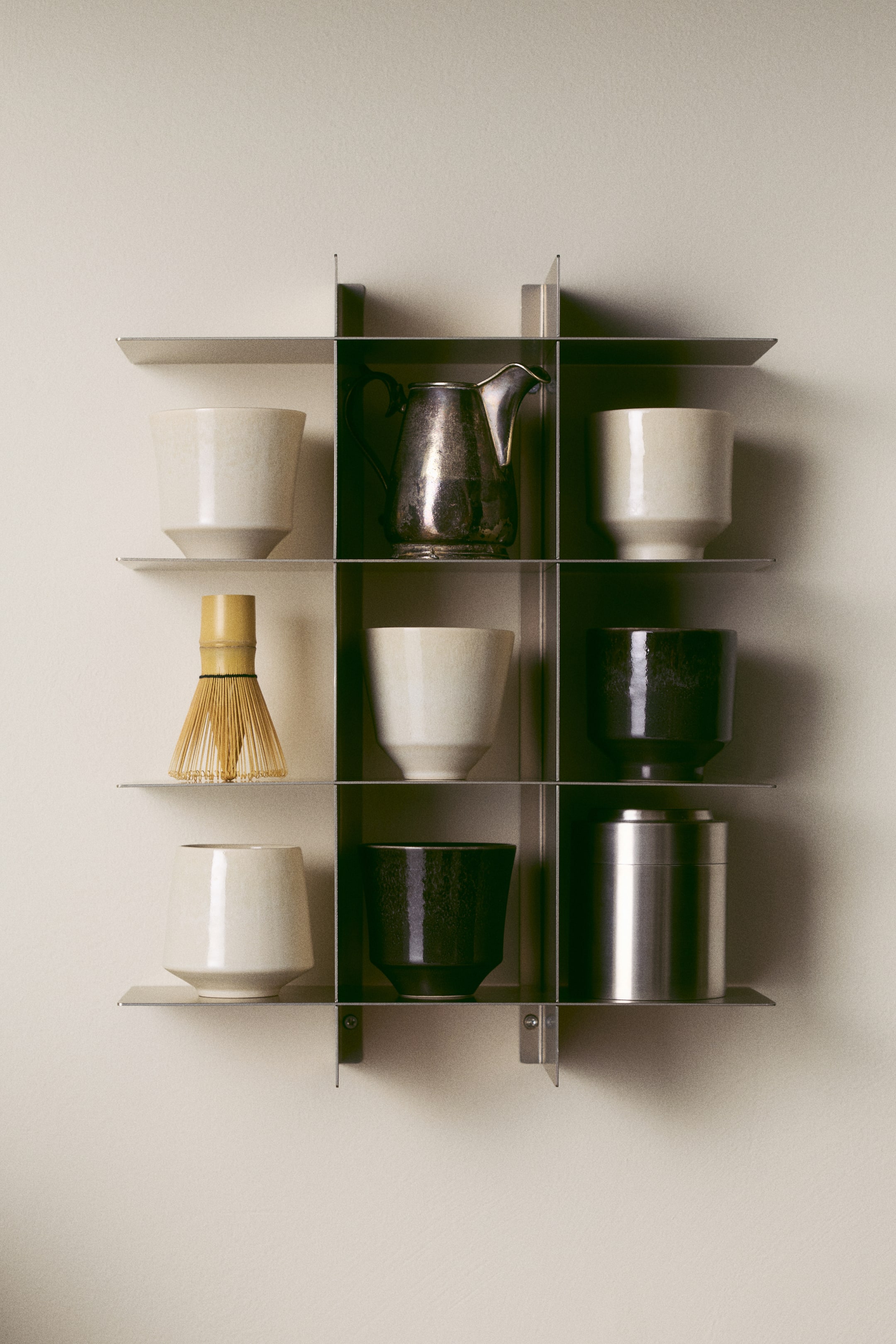 View larger image: A front-facing view of a wall shelf in brushed metal featuring an open grid design with multiple shelves and compartments. The shelf displays various decorative items, including ceramic cups, a small metal pitcher, a bamboo whisk, and a metal canister, highlighting its storage use.