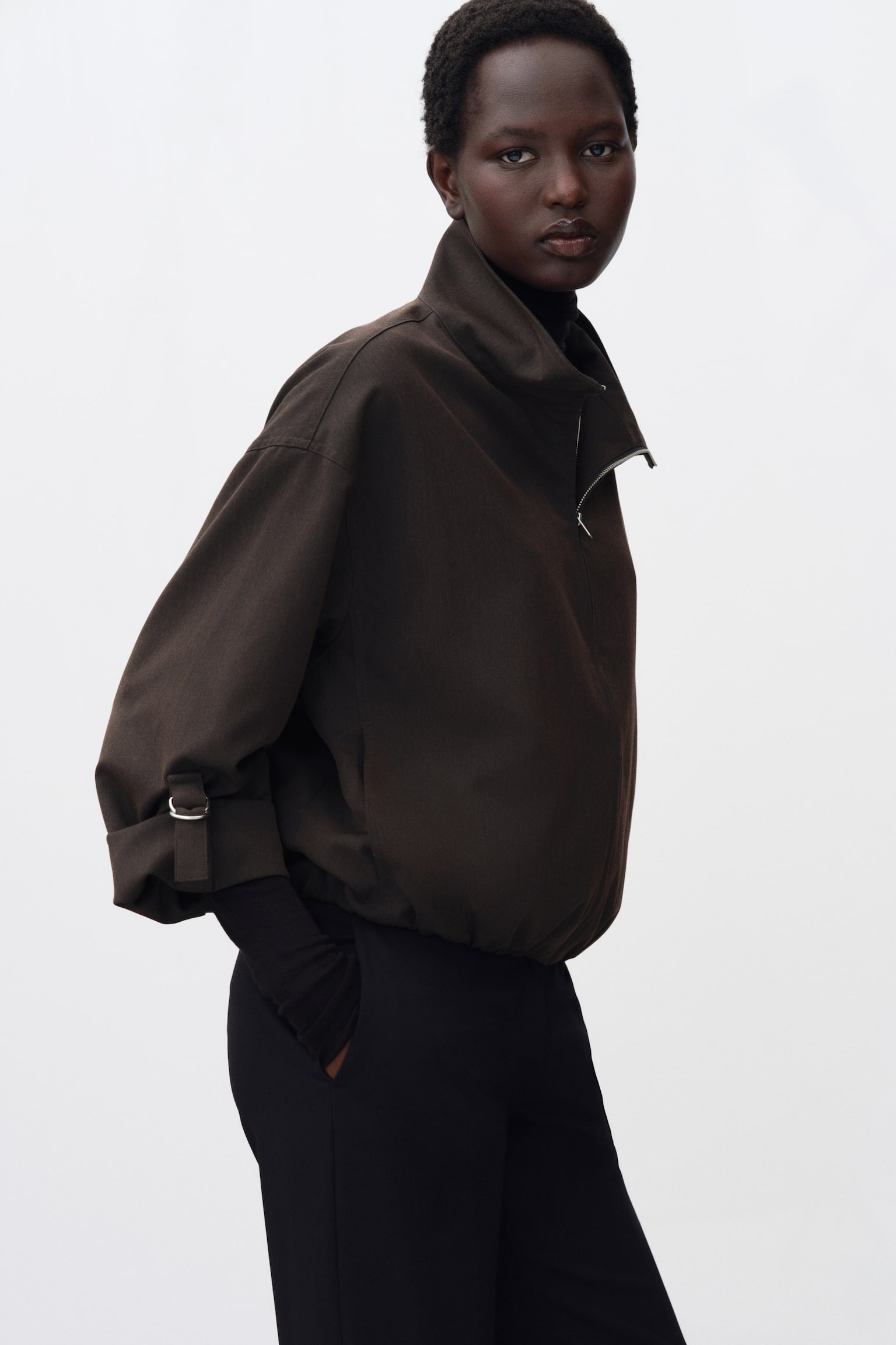 A short, oversized jacket in dark brown is being worn by the model, featuring a funnel collar with a partially unzipped front, dropped shoulders, and rolled-up cuffs secured with metal D-rings. The jacket is layered over a black high-neck top and black trousers. - 1