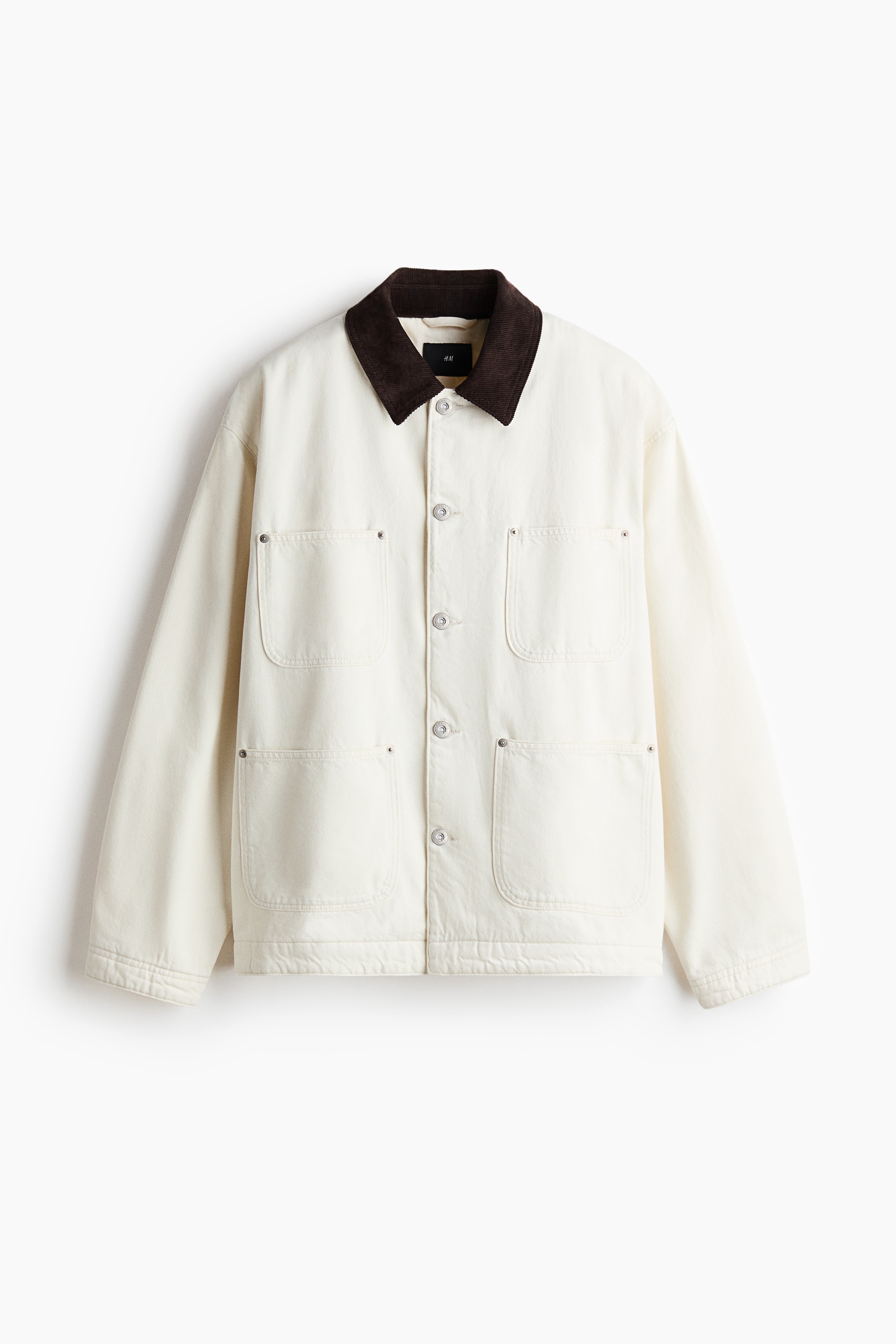 An off-white denim chore jacket featuring a contrasting dark brown corduroy collar is worn open by the model. The jacket has patch pockets on the chest and lower front, and is layered over a dark blue T-shirt and blue jeans.