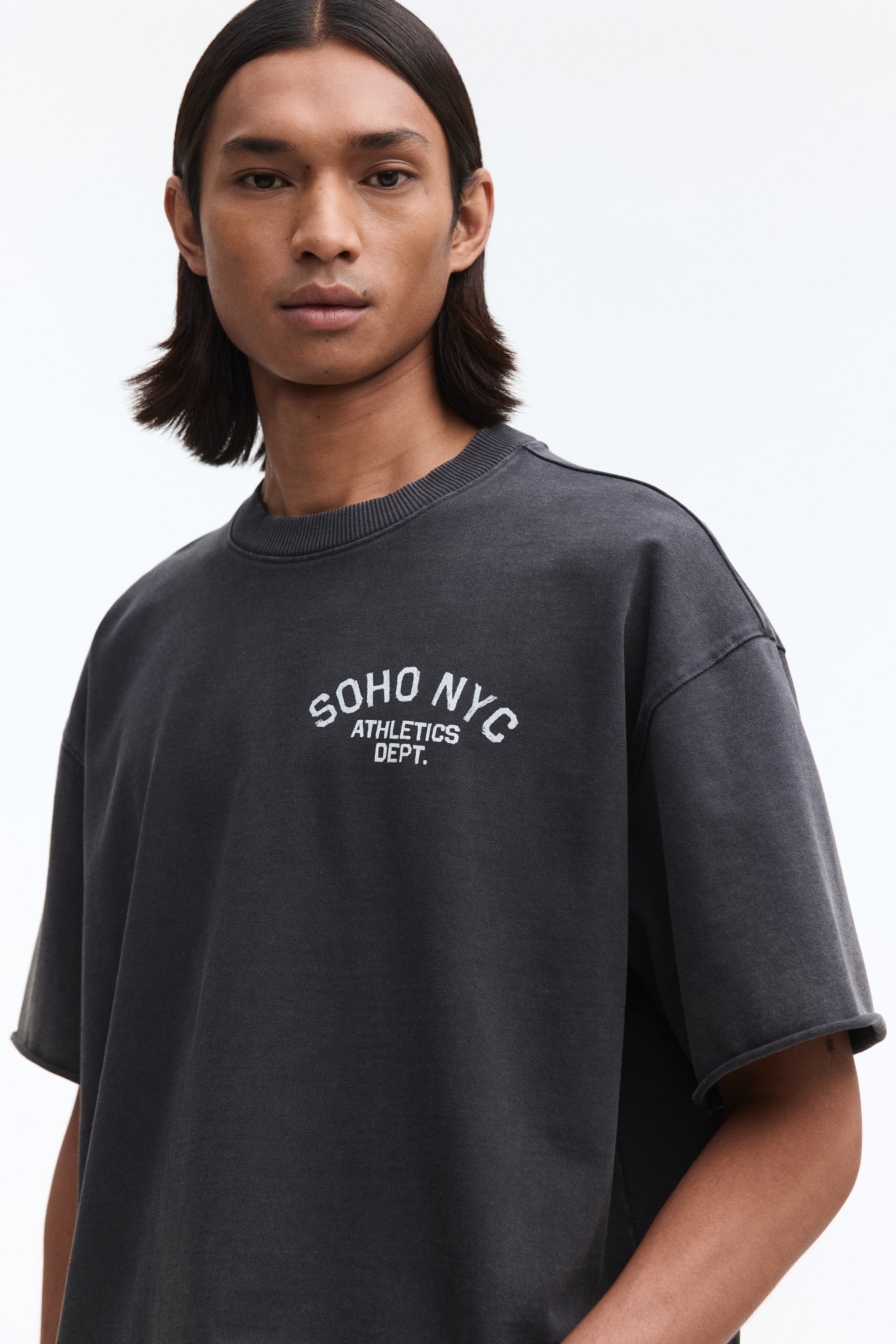 View larger image: A washed dark grey short-sleeved t-shirt featuring a ribbed crew neck and raw-edge sleeve hems. Its front displays "SOHO NYC ATHLETICS DEPT." in distressed white text.