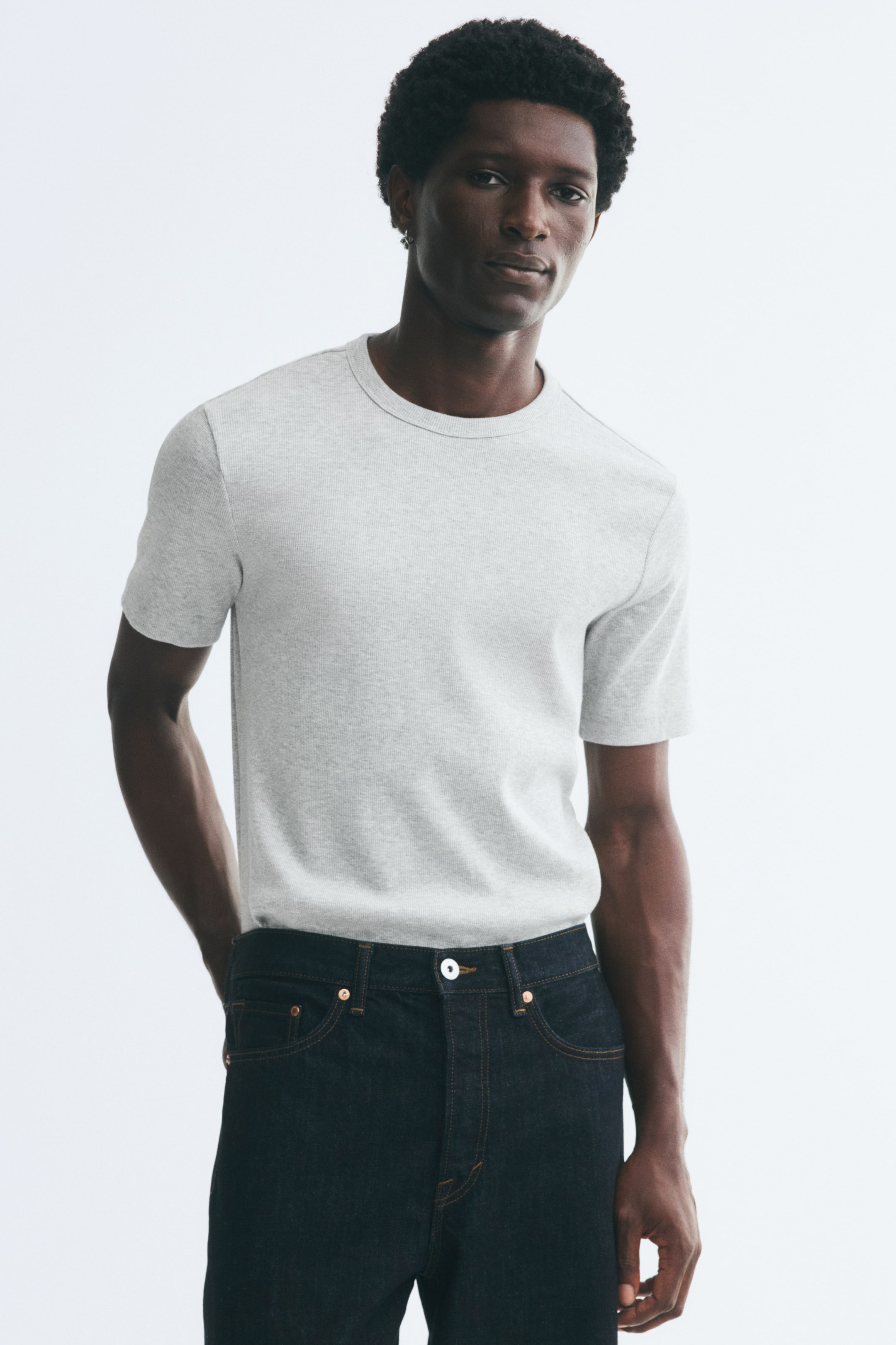 Agrandir l'image: Un t-shirt en jersey de coton côtelé gris clair est porté par le mannequin, avec une coupe ajustée, un col rond et des manches courtes. Le t-shirt est rentré dans un jean en denim foncé.