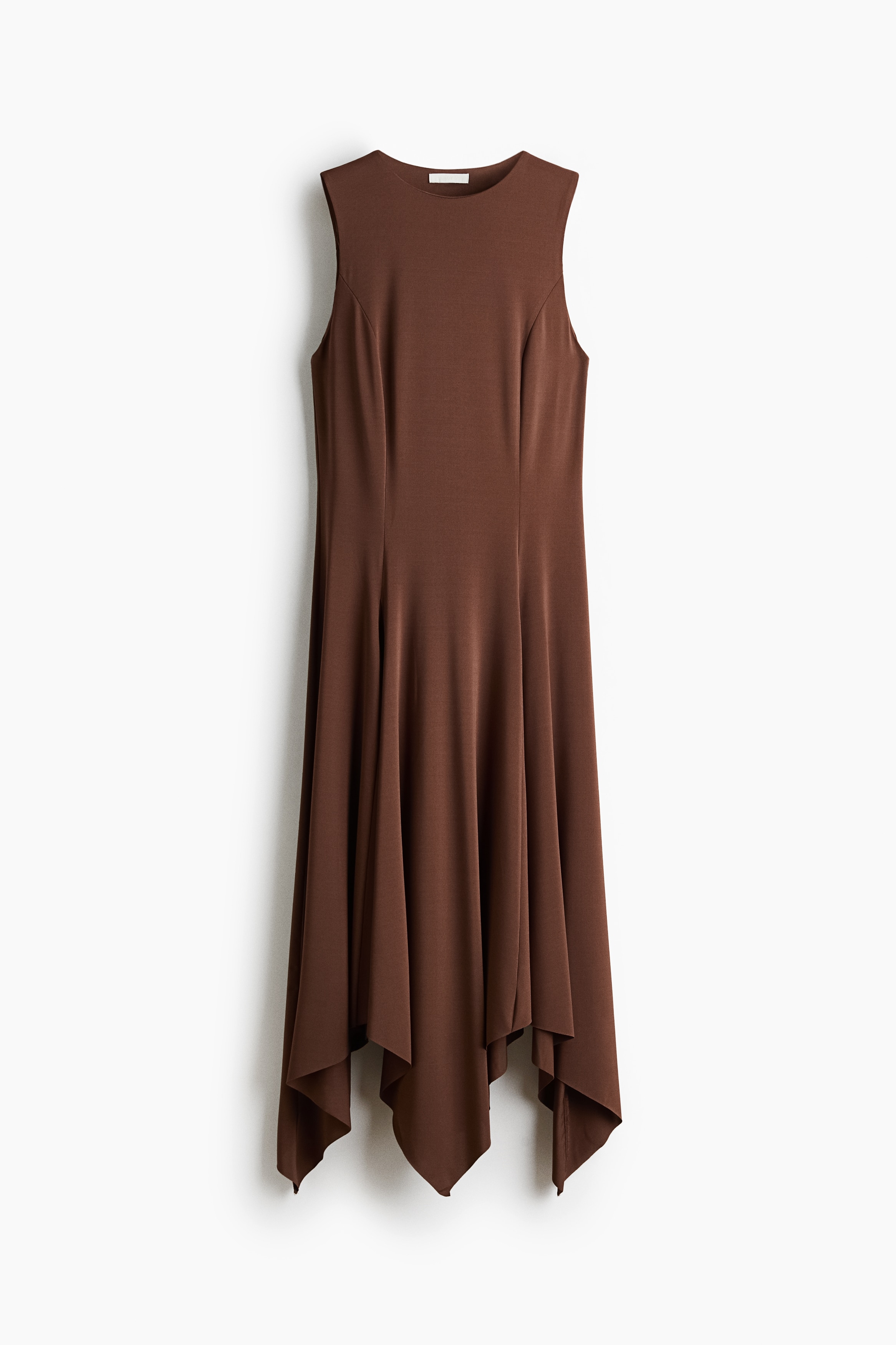 A brown, sleeveless, slim-fit dress with a round neckline is being worn by the model. The dress features princess seams that open into a flared skirt with godet pleats and an asymmetrical, handkerchief hem, styled with a black handbag held under the arm and black flat sandals.