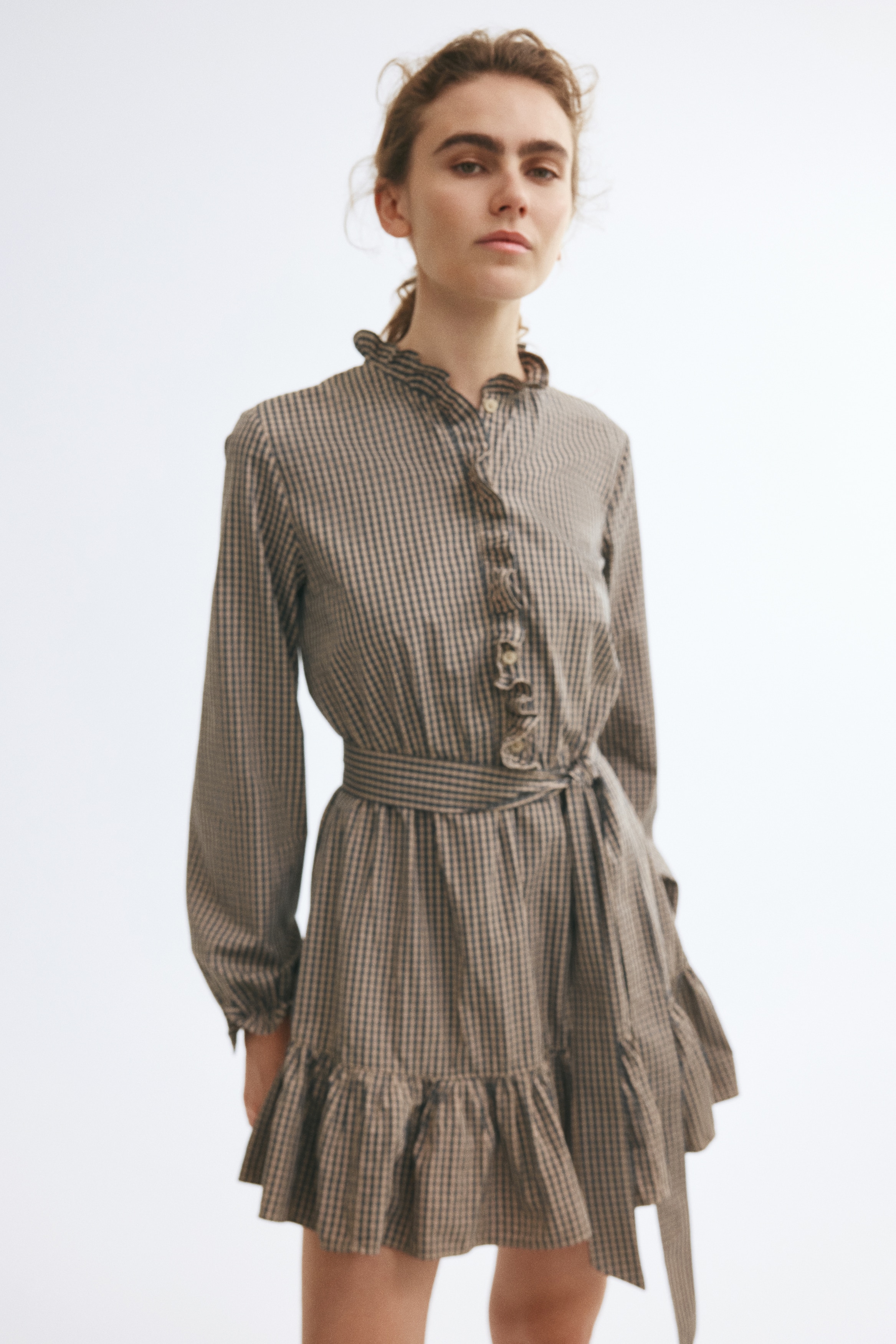 A long-sleeved gingham mini dress in brown and black is shown from the front, featuring a ruffled high collar and a matching ruffled front placket. The dress is cinched at the waist with a matching fabric tie belt and features long sleeves with ruffled cuffs and a ruffled hem.