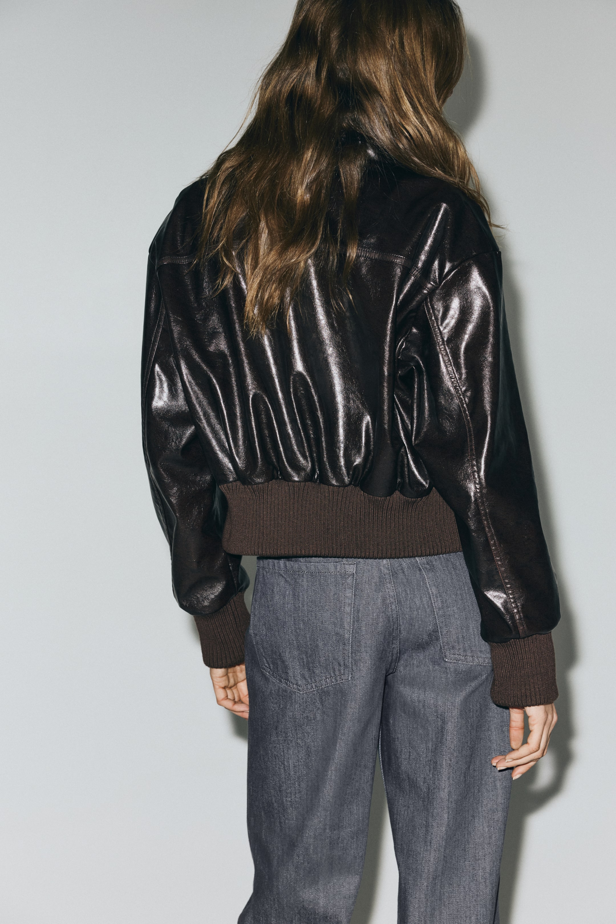 Agrandir l'image: Une veste bomber marron foncé et brillante est visible de dos, avec des poignets et un ourlet côtelés. Elle est portée par-dessus un pantalon gris, qui montre des poches arrière visibles.