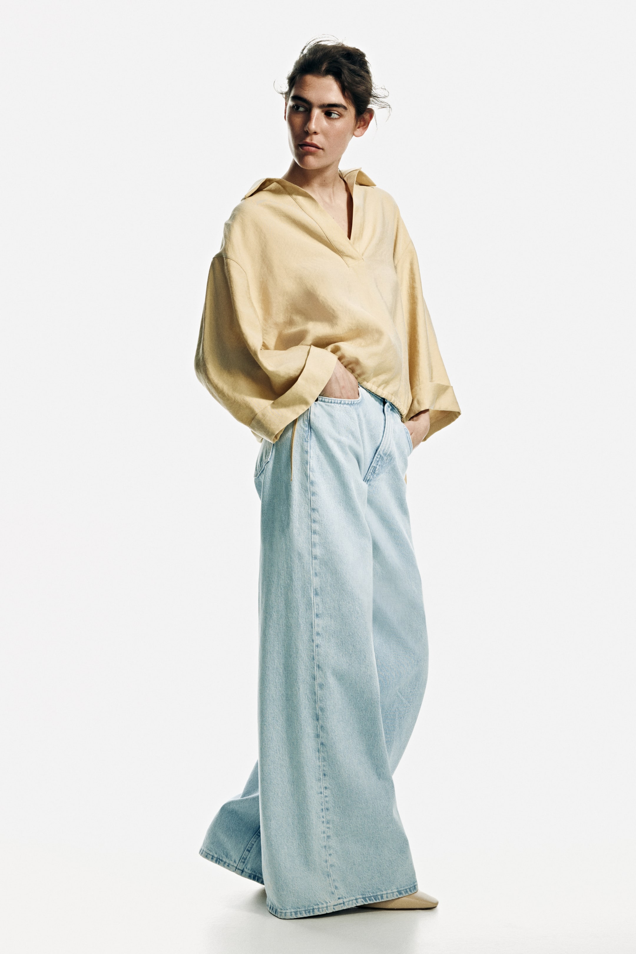 Grösseres Bild ansehen: Ein hellgelbes, übergroßes Hemd mit Kragen, V-Ausschnitt, weiten Ärmelbündchen und gerafftem Saum wird in einer Dreiviertel-Frontansicht präsentiert. Es wird kombiniert mit hellblauen, hoch taillierten Jeans mit weitem Bein, die am Boden aufliegen.