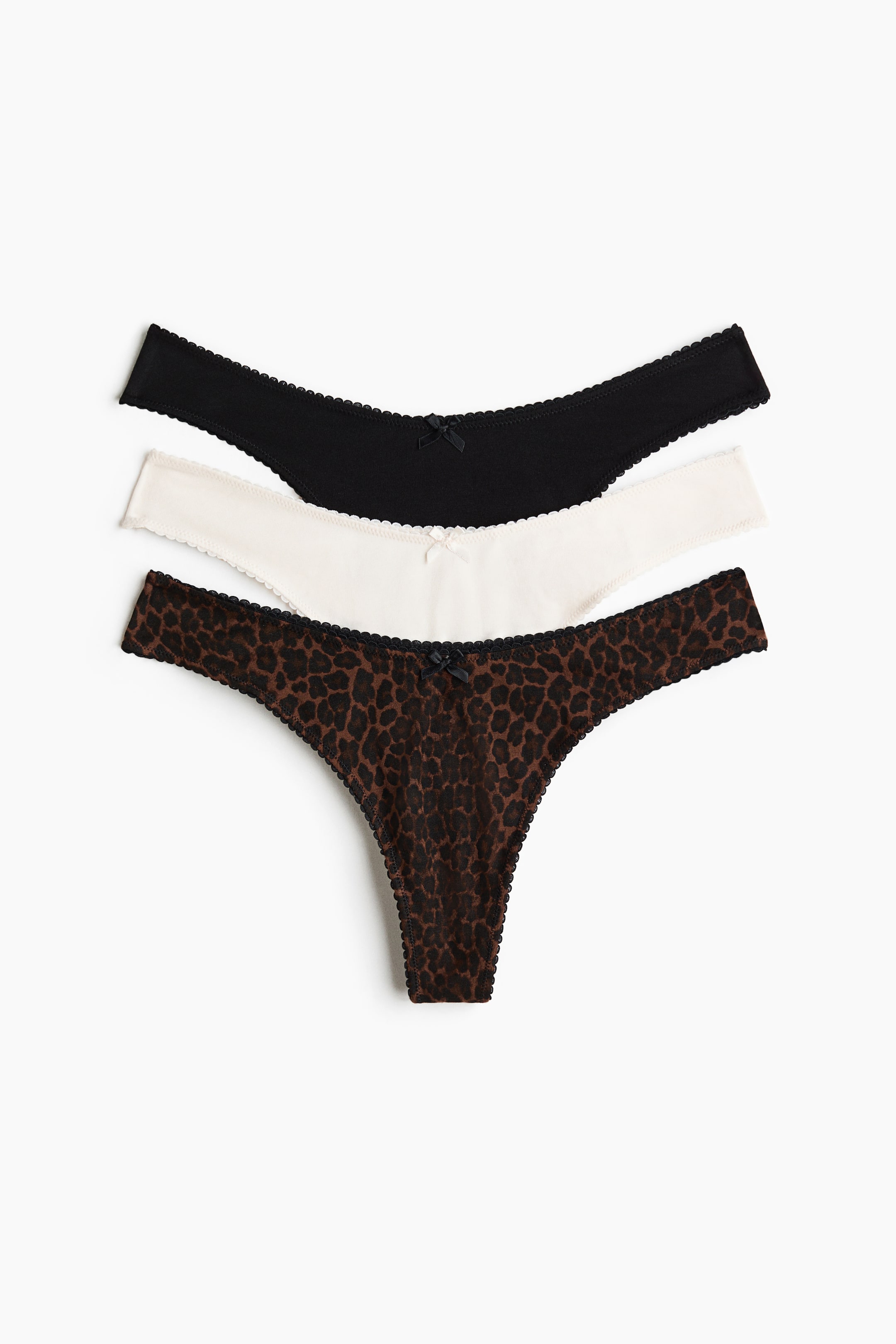Ver imagen más grande: Se muestran tres braguitas tipo tanga de punto de algodón, colocadas en plano, apiladas y ligeramente desfasadas. El conjunto incluye una braguita negra, una en blanco roto y una con estampado de leopardo marrón, cada una con cintura baja, un pequeño lazo en la parte delantera y un acabado ondulado en los bordes.