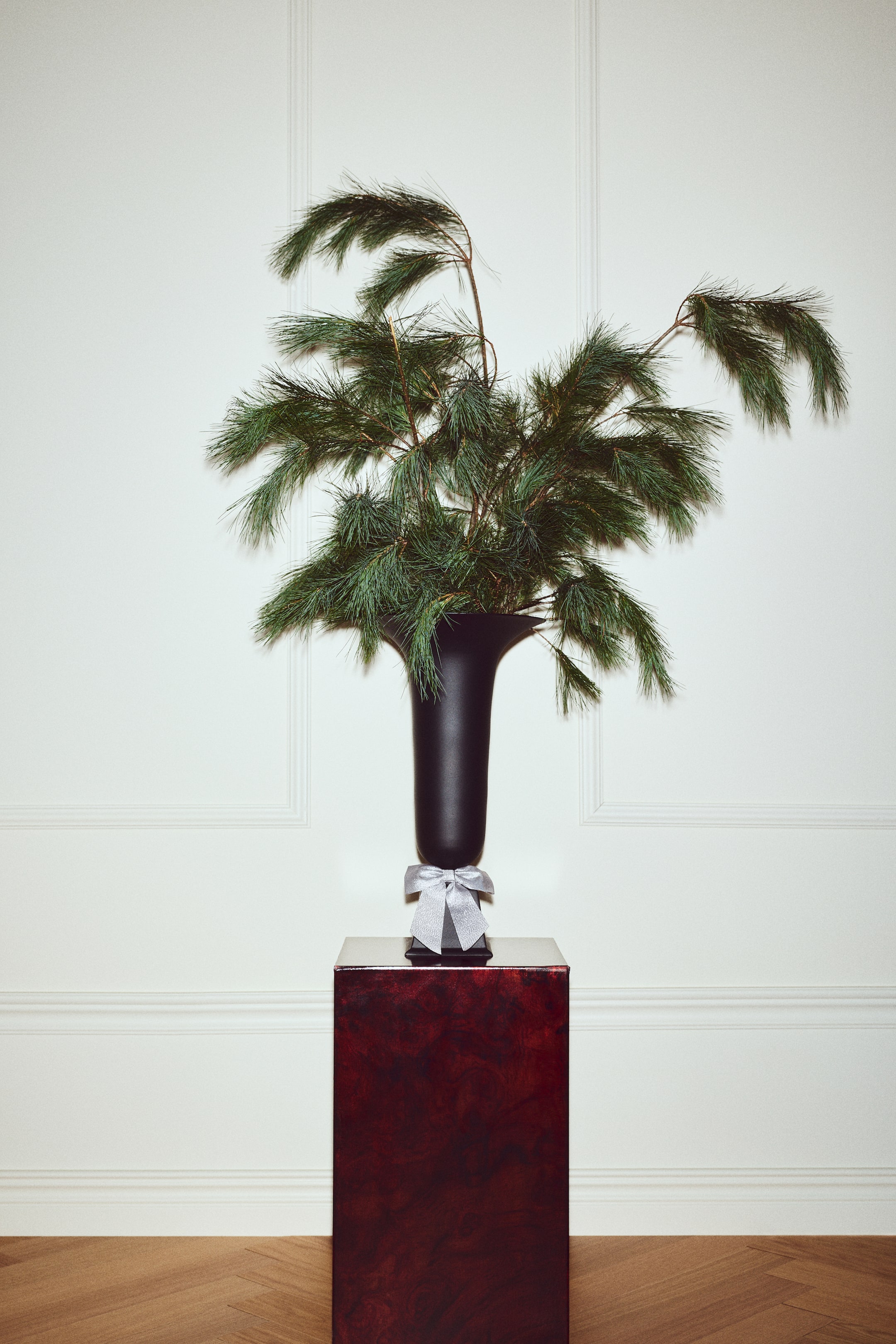 Agrandir l'image: Une décoration en velours gris clair en forme de nœud est enroulée autour de la base d’un vase noir, orientée vers l’avant. La décoration met en valeur des boucles structurées et deux larges pans inclinés qui soulignent sa texture douce et moelleuse.