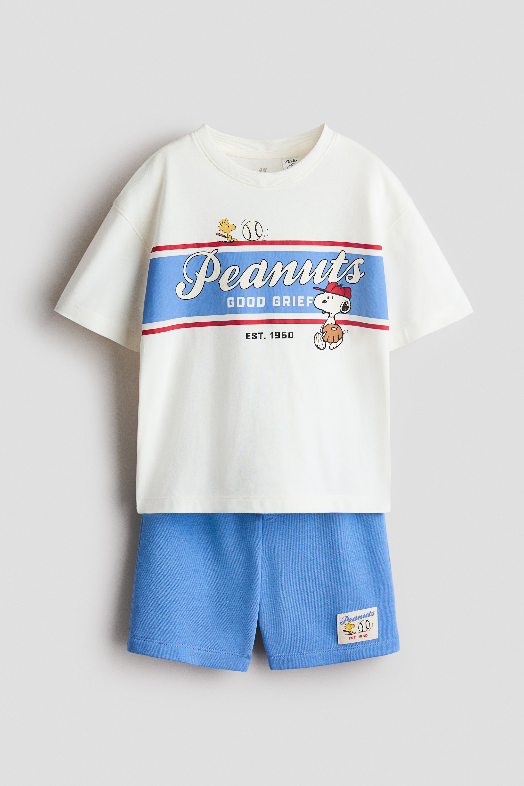 View larger image: A two-piece clothing set is shown front-facing, consisting of a cream-colored cotton T-shirt and blue sweat shorts. The T-shirt features a round ribbed neckline, dropped shoulders, and a baseball-themed "Peanuts" graphic with Snoopy and Woodstock across the chest. The matching blue shorts feature a small rectangular graphic patch on the lower left hem.
