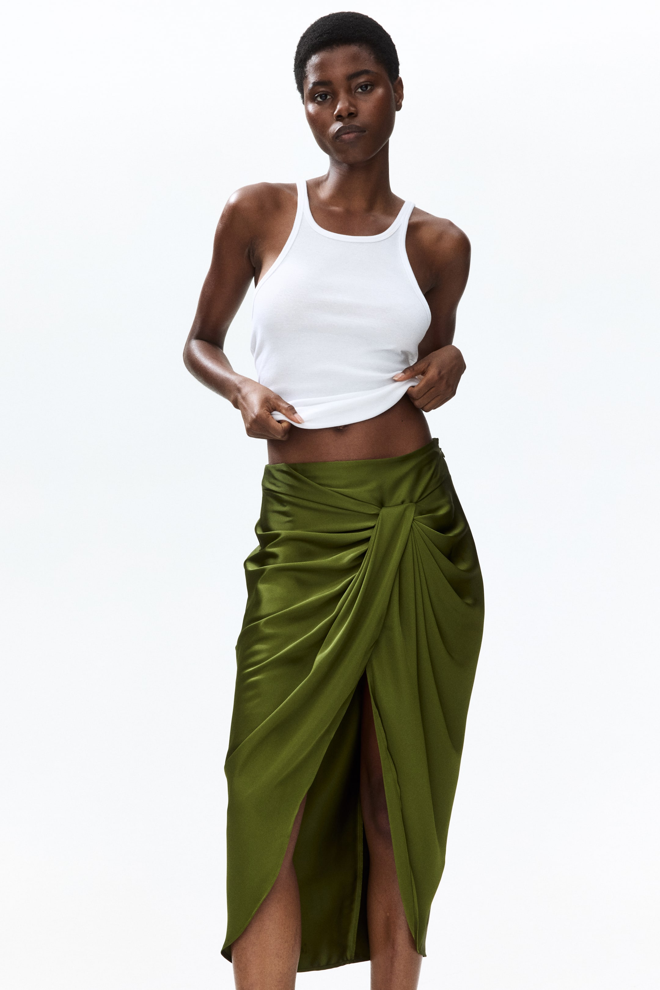 View larger image: An olive green satin midi skirt showcasing a twisted front detail that forms a high slit, with the fabric flowing elegantly to an asymmetrical hem.