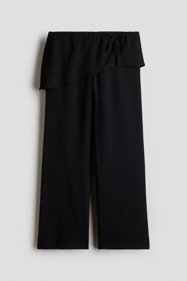 A pair of black wide-leg trousers is shown from the front, featuring a textured, crinkled fabric and an integrated asymmetrical wrap overlay at the waist. The overlay is draping diagonally across the hips and securing with a small tie detail on the upper right side. - 1