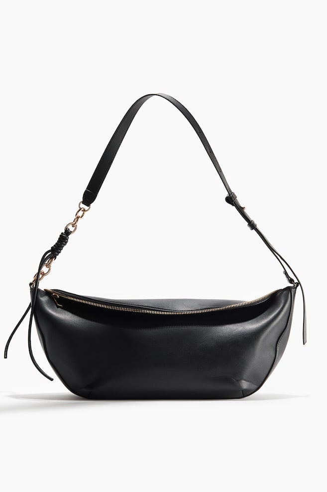 The black, crescent-shaped handbag showcases a smooth, supple material with a gold-toned zipper closure along its top. An adjustable shoulder strap connects to the bag with gold-toned chain links, featuring a wrapped detail on one side, as the bag is presented from a slightly elevated front angle. - 1