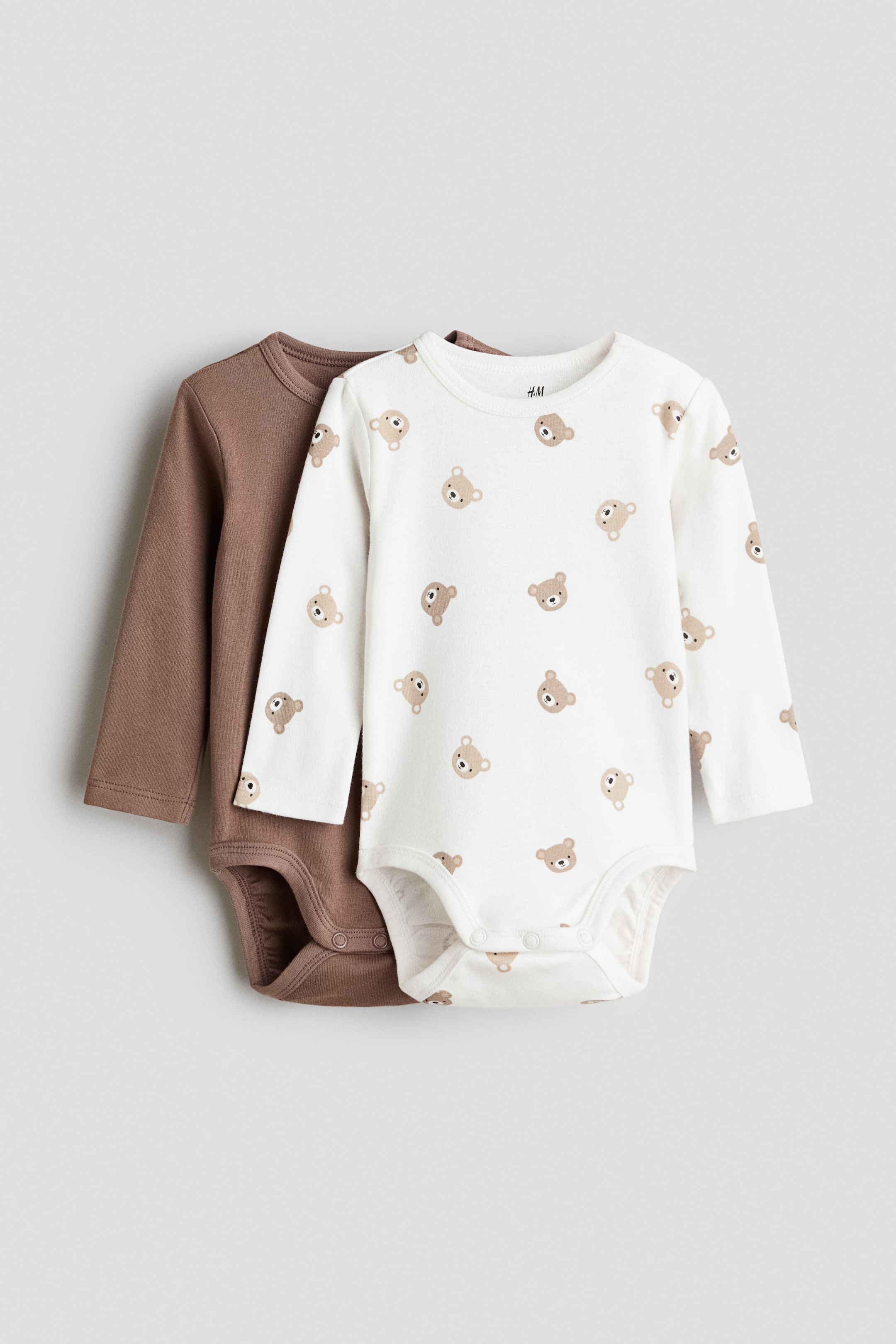 View larger image: Two bodysuits are displayed with their front sides facing up, lying flat. The front bodysuit is off-white, adorned with a repeating pattern of small brown bear heads, featuring long sleeves and snap buttons at the crotch. Behind it, a solid brown bodysuit with long sleeves is subtly visible, adding a touch of contrast.