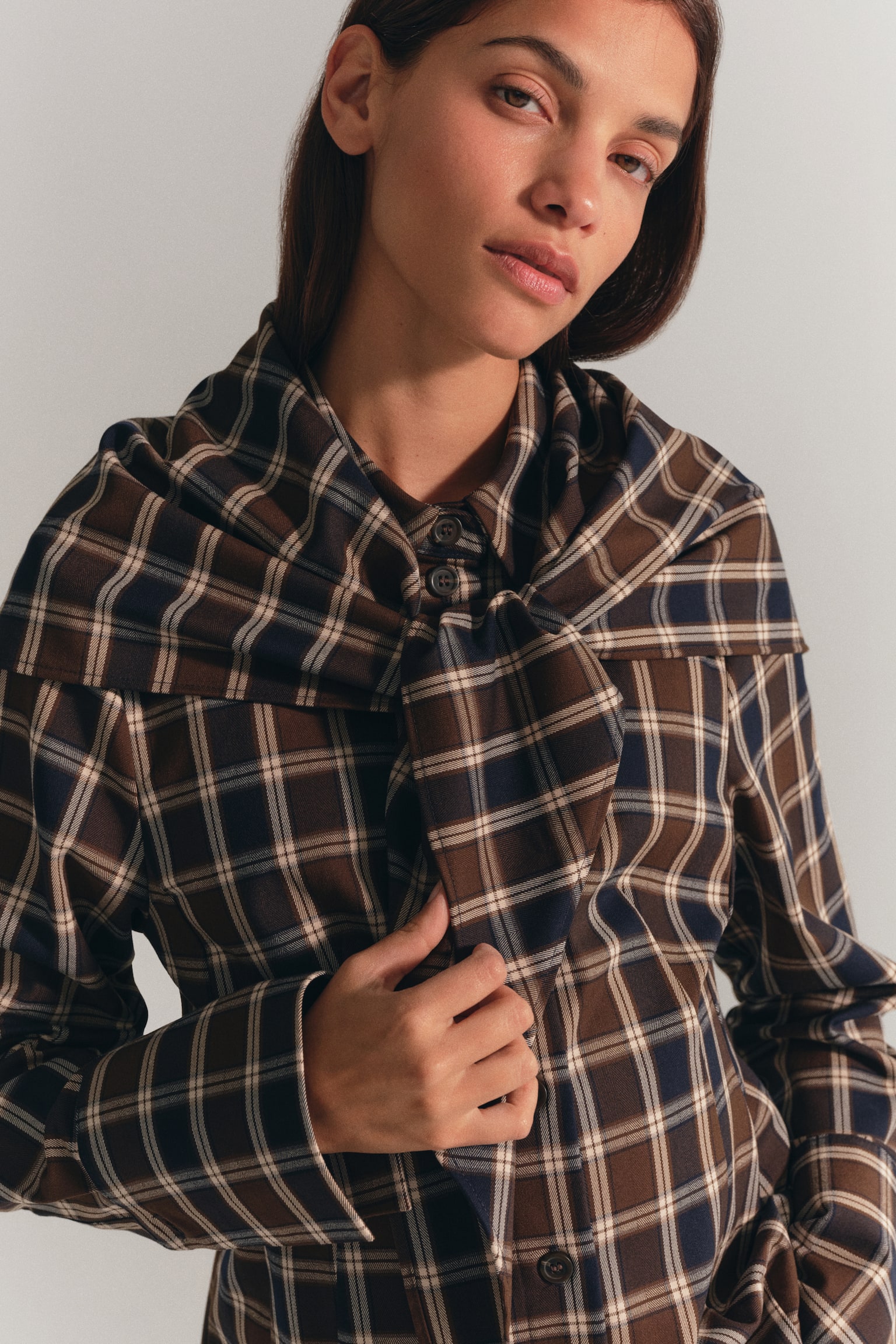 A plaid twill shirt in brown, navy, and white is worn by the model, who is holding one end of the matching scarf draped over the shoulders. The shirt features a button-down front, a pointed collar, and extra-long sleeves with wide cuffs. - 1