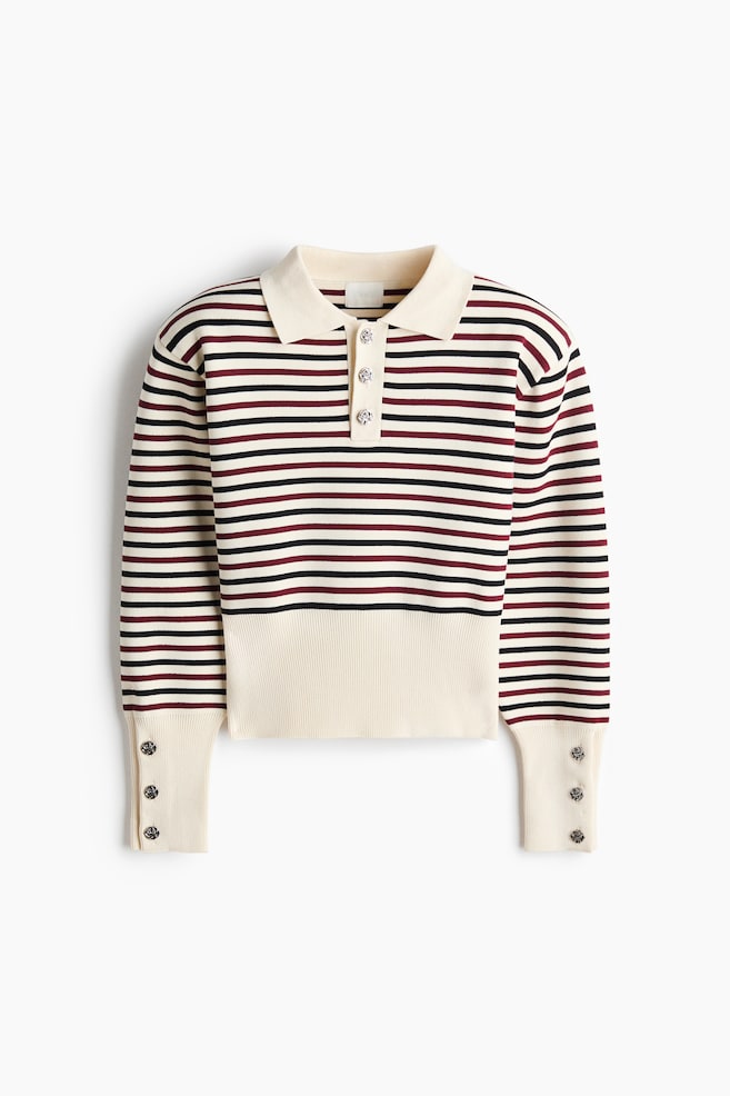 A short, fine-knit polo shirt is shown from the front, featuring a cream base with horizontal black and burgundy stripes. It has a classic collar, a three-button placket with decorative metallic buttons, and structured shoulders. The long sleeves end in wide, buttoned ribbed cuffs that coordinate with a wide, solid cream ribbed hem. - 1