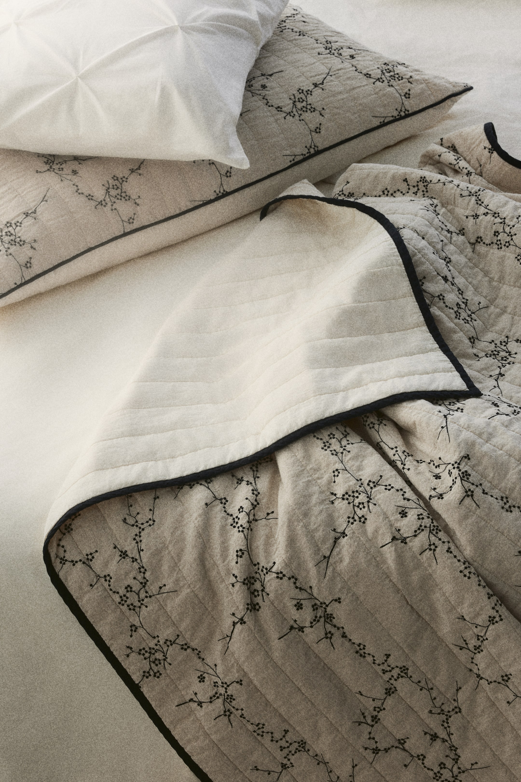 Ver imagen más grande: Un juego de ropa de cama acolchada en beige claro presenta una manta y una funda de almohada a juego, ambas con un delicado estampado floral negro y un ribete negro a lo largo de sus bordes. La manta está doblada, revelando su reverso acolchado en crema con costuras horizontales.