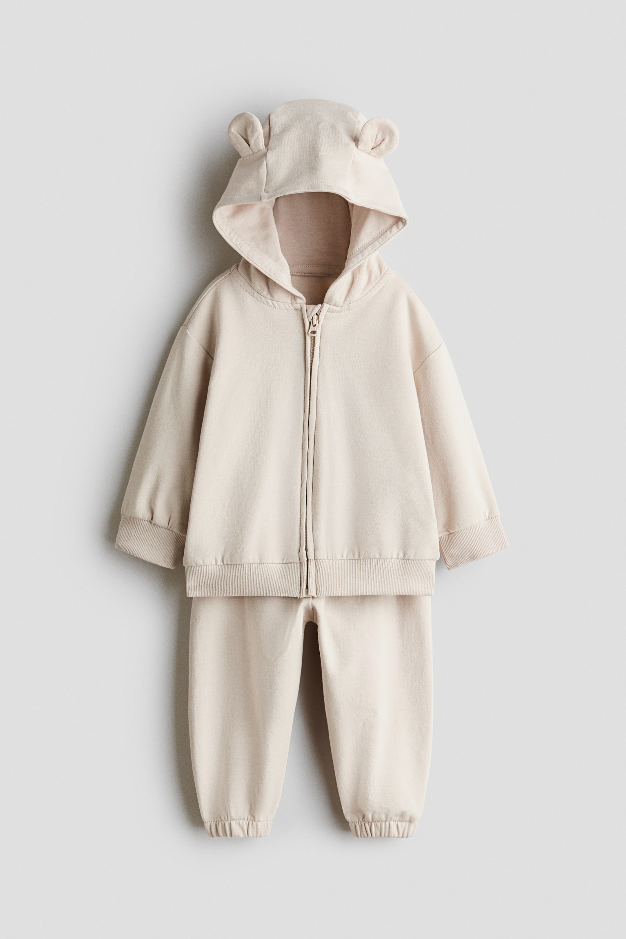 View larger image: A front view of a light beige two-piece sweat set featuring a zip-up hoodie with small fabric ears on the hood, dropped shoulders, a front zipper, and ribbed cuffs and hem. The matching track pants are displayed below, featuring elasticised ankle cuffs.