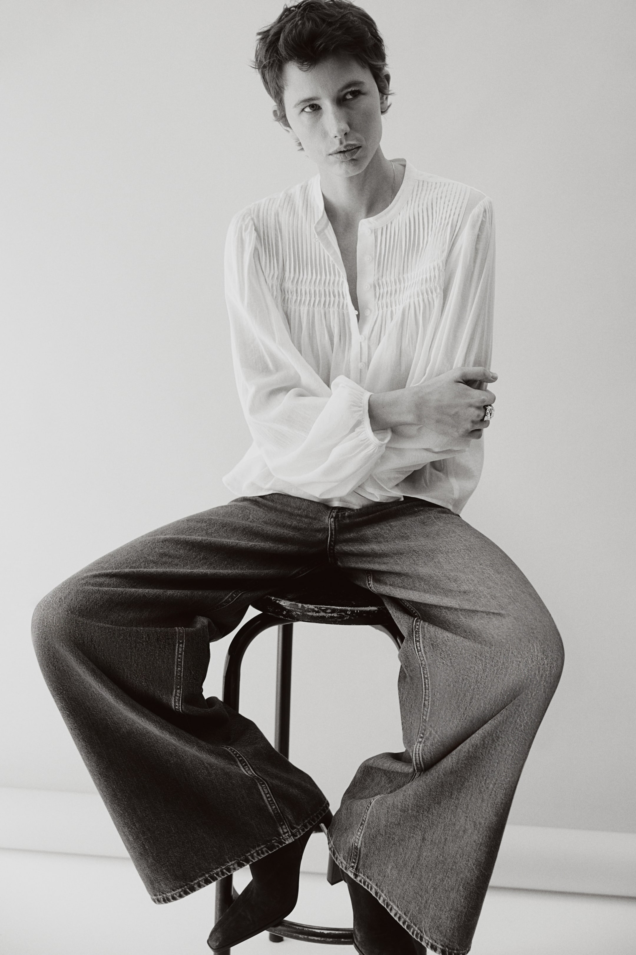 Ingrandisci l'immagine: Una blusa chiara presenta dettagli pintuck sul corpetto anteriore e maniche voluminose raccolte, ed è abbinata a jeans scuri a gamba larga dal relaxed fit che si adagiano sui piedi.