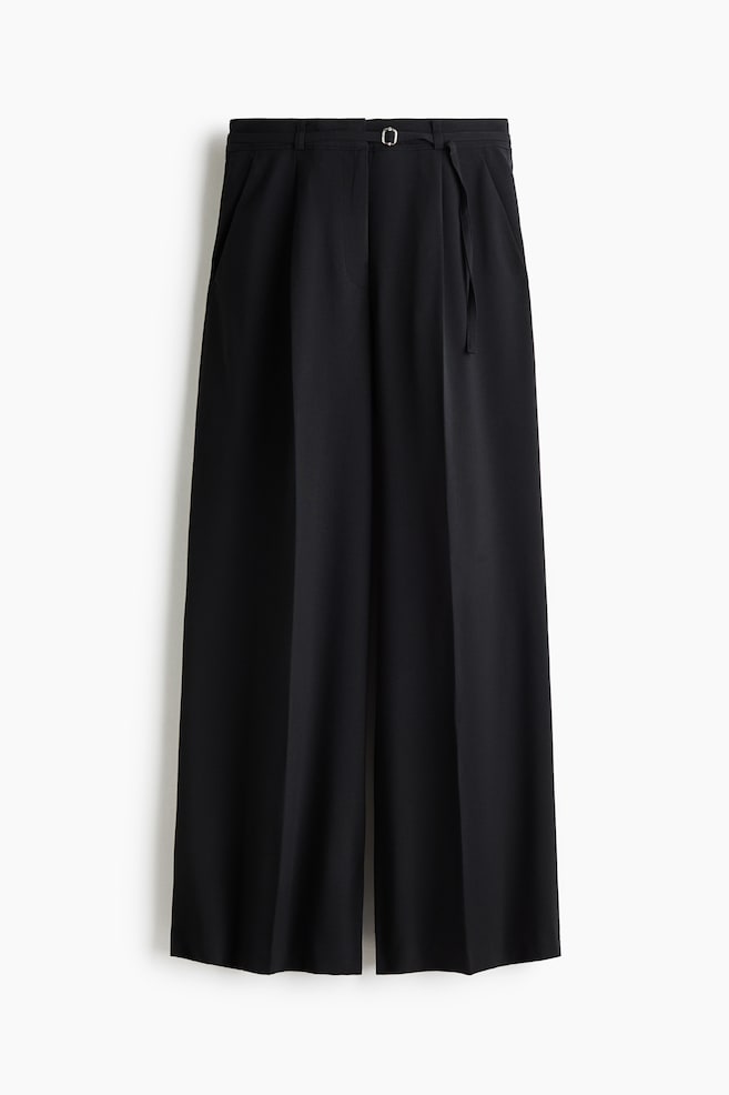 A pair of loose-fit tailored black trousers is hanging at a front-facing angle, showing a high waist with a narrow adjustable belt and diagonal side pockets. The wide legs fall straight down with prominent pressed creases running down the centre of each leg. - 1
